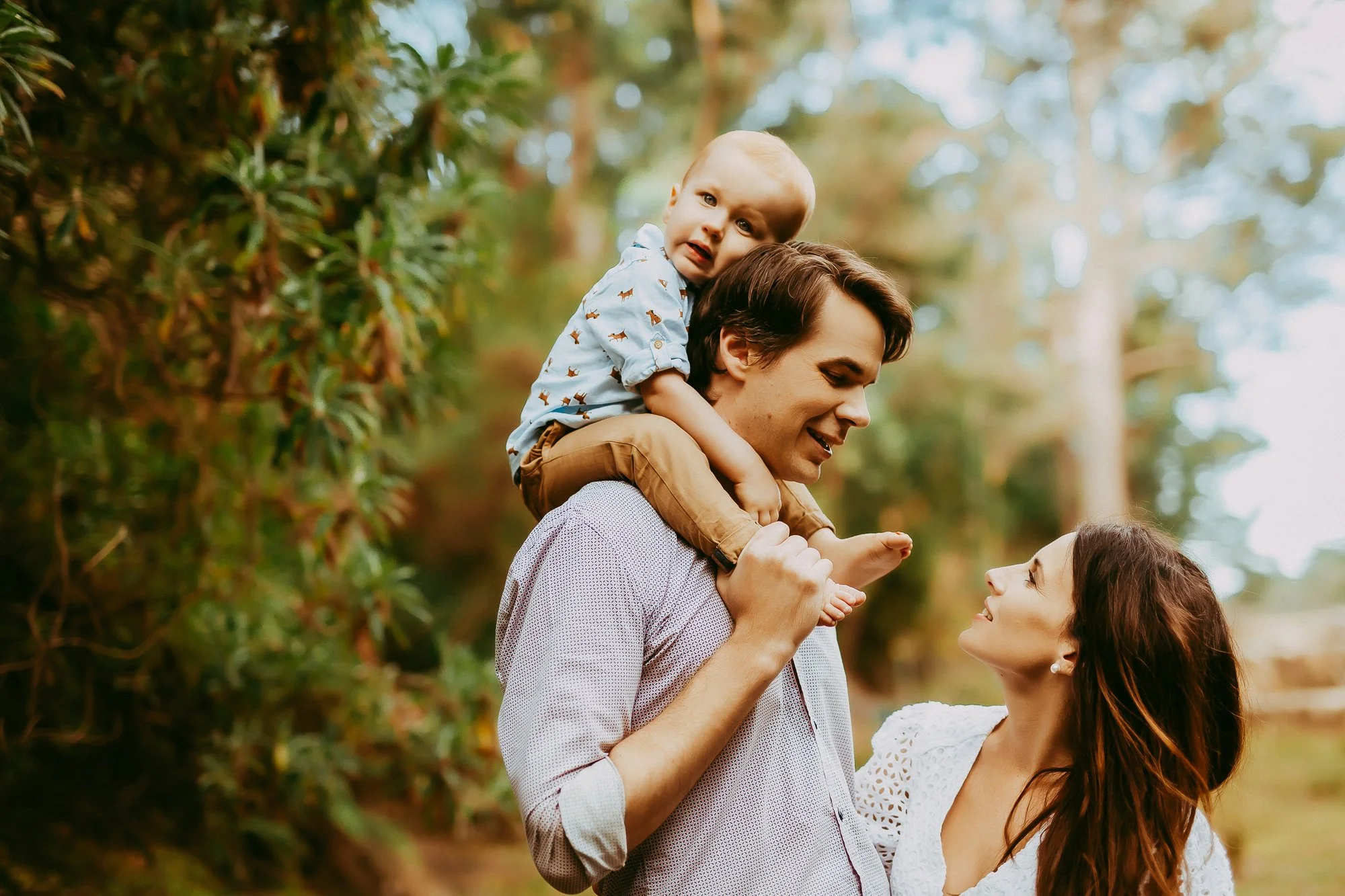 A happy family of three enjoying time outdoors in a forested area, with the father carrying their young son on his shoulders, and the mother smiling at them.