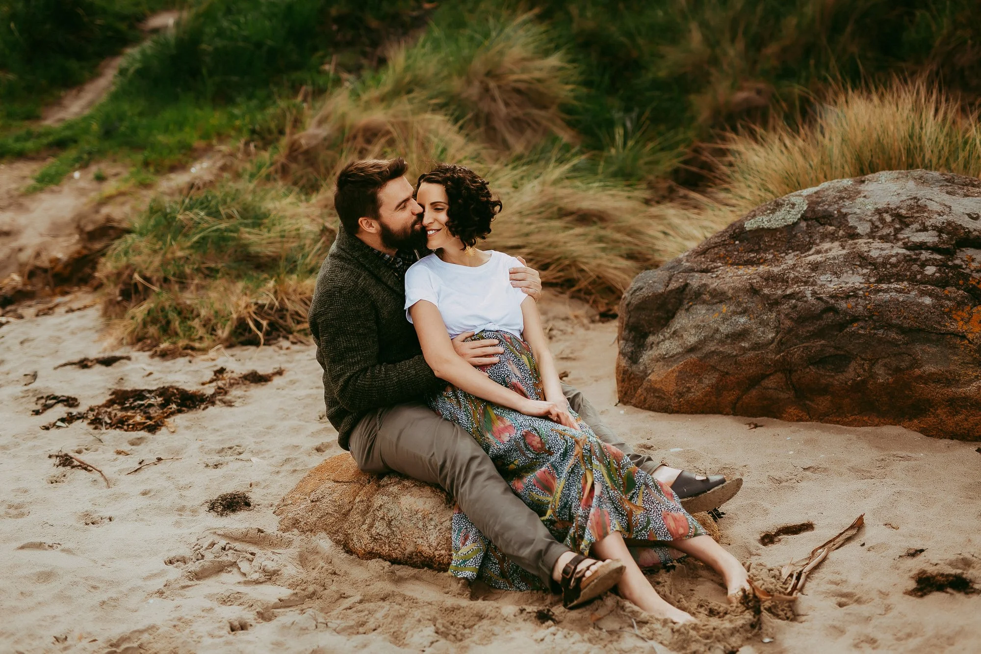 A couple sitting on a large rock on a sandy beach, embracing each other and smiling, with grassy dunes and rocks in the background.