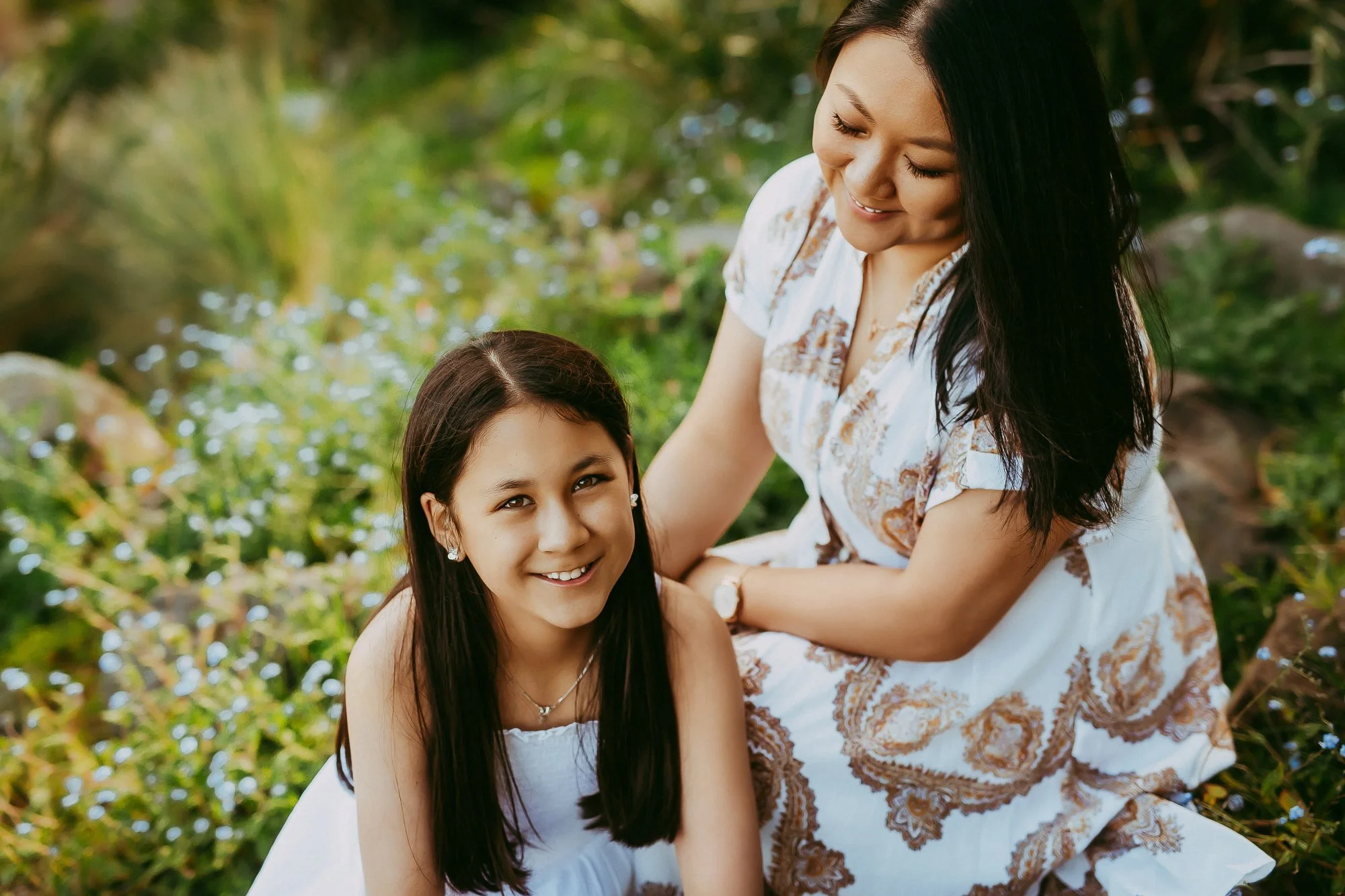 A woman and a young girl sitting outdoors among greenery, smiling and looking at the camera.