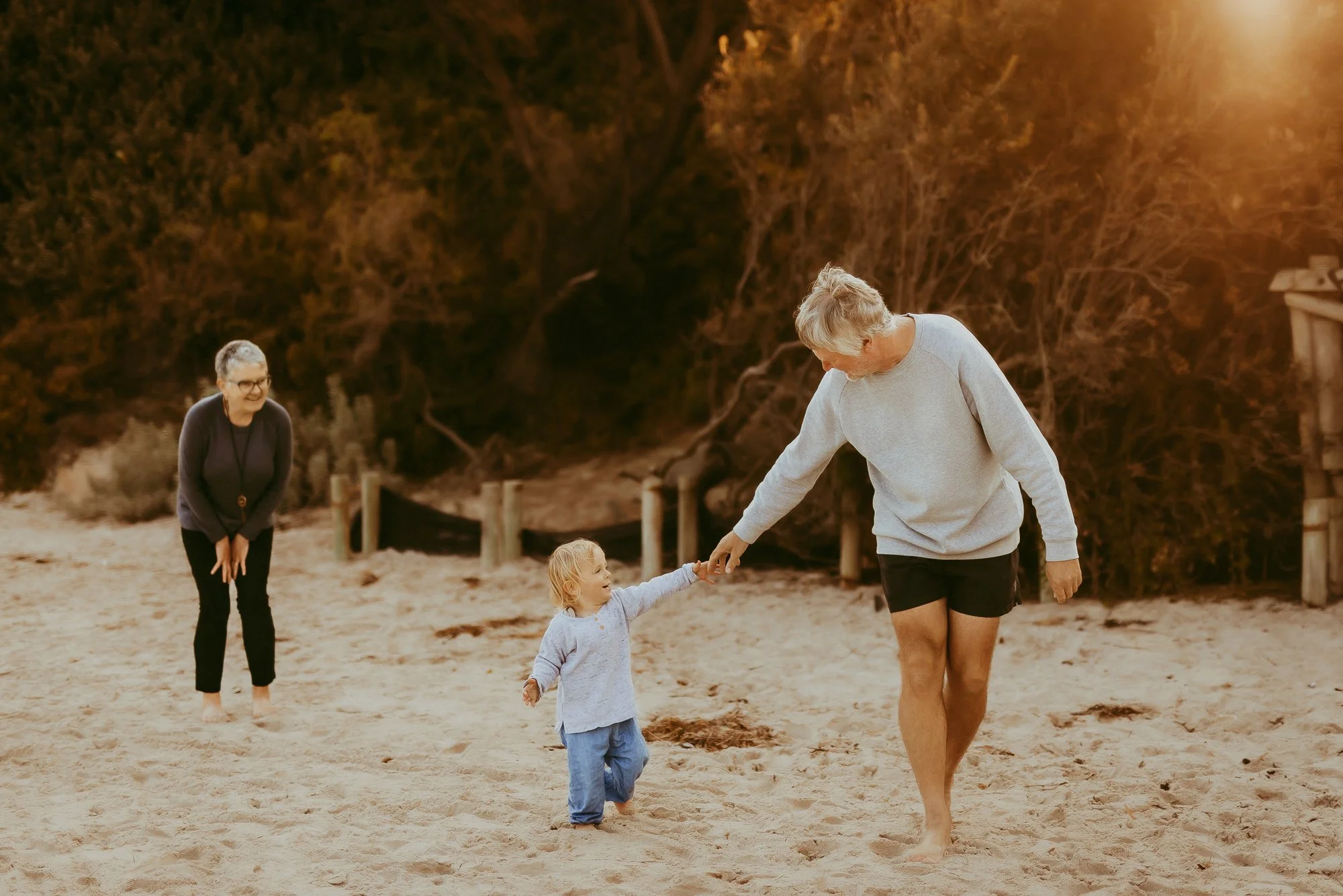 Three people on a sandy beach, with a young child holding hands with an elderly woman, and another woman smiling nearby, all enjoying a sunny day.