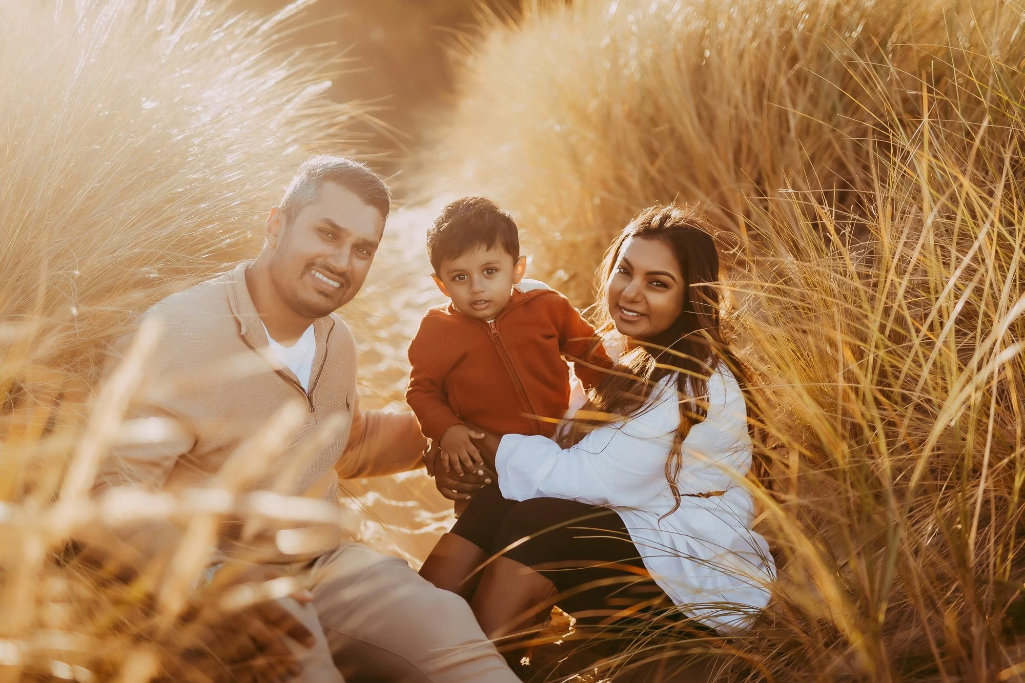 A family of three, a man, woman, and young boy, sitting in a field of tall, golden grass during sunset, smiling at the camera. Golden hour family photosession at Seven Mile Beach Tasmania by Ulla.