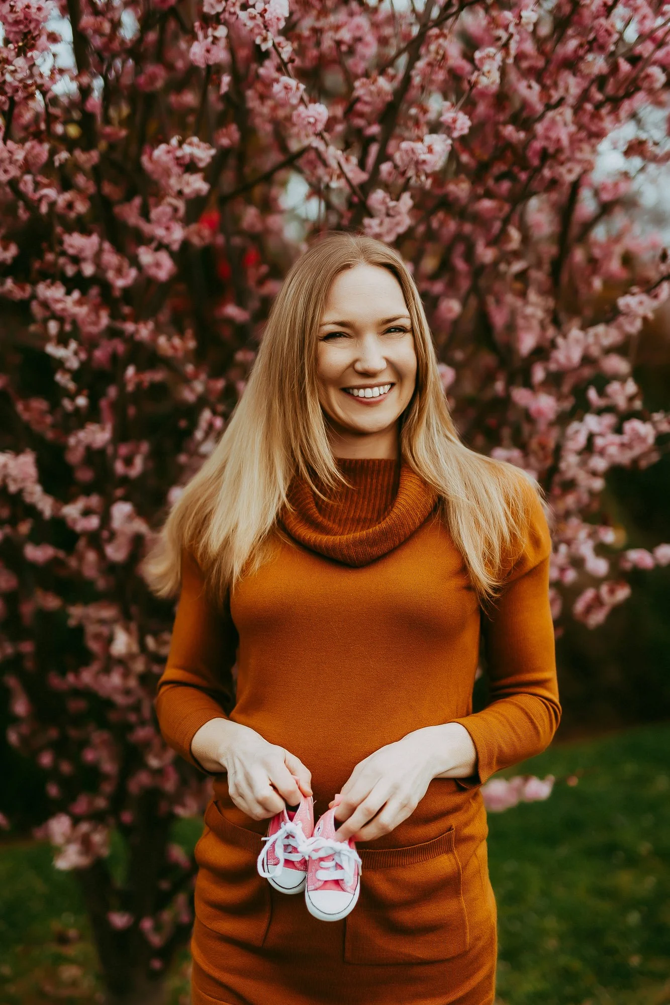 A young woman with blonde hair smiling and holding small pink and white sneakers, standing outdoors in front of a blooming pink cherry blossom tree.