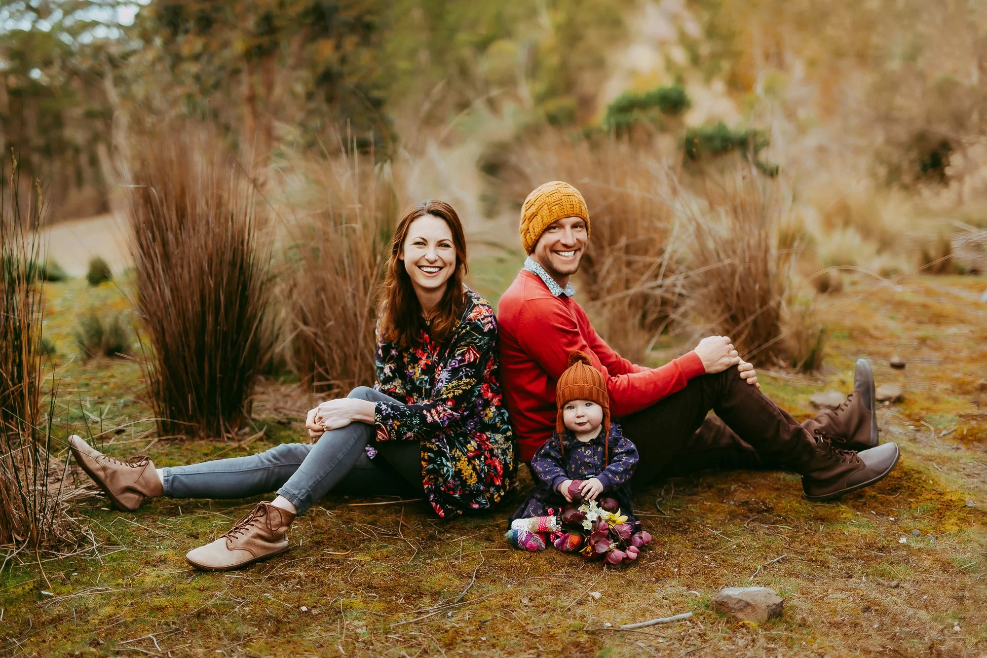 Family of three including a woman, a man, and a baby sitting outdoors on autumn ground near tall brown plants, smiling at the camera. Hobart area family photosession.