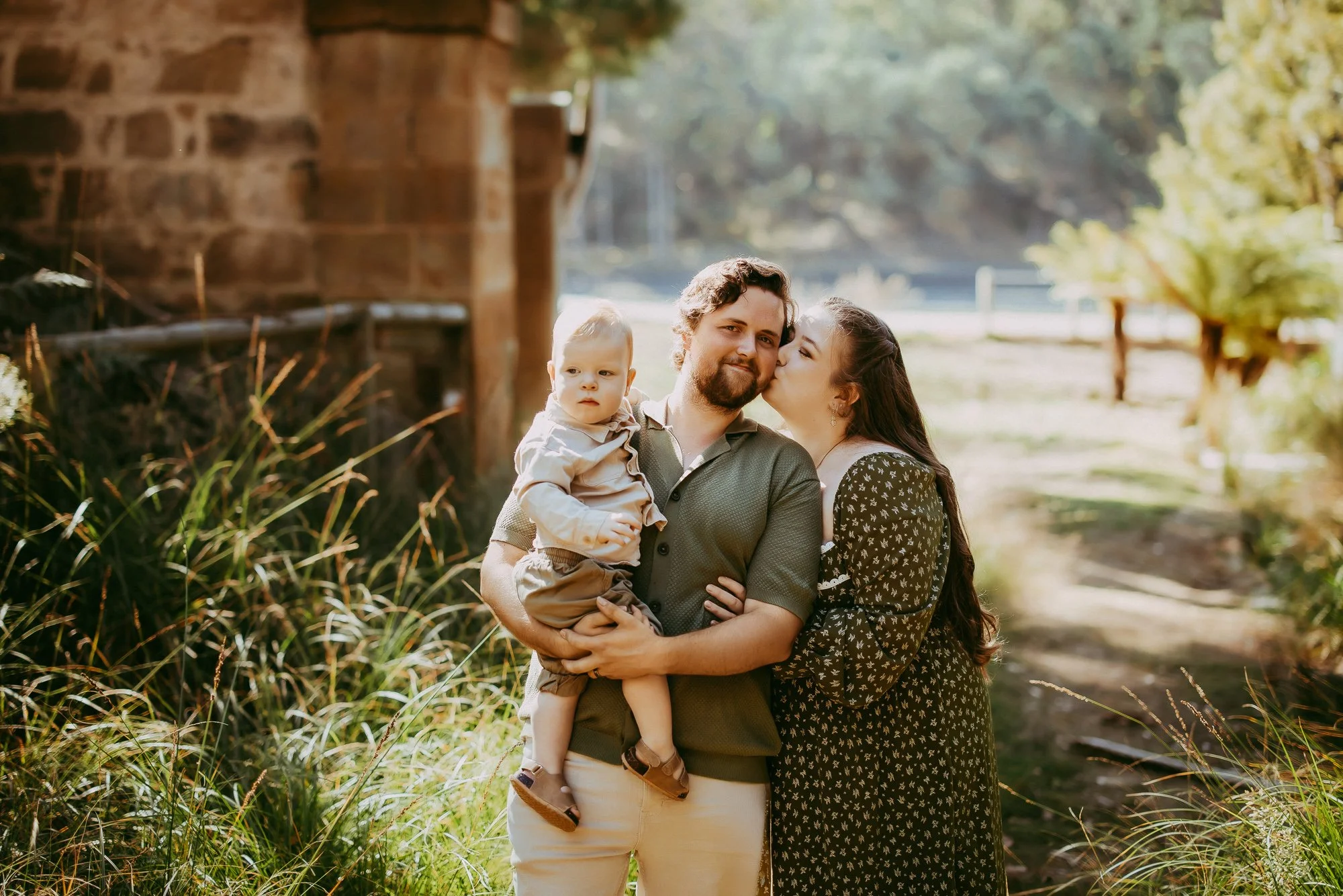 A family of three outdoors in warm sunlight, with a father holding a young son and a mother kissing his cheek, near a rustic building and greenery. Budget friendly family photographer at Waterworks Reserve.