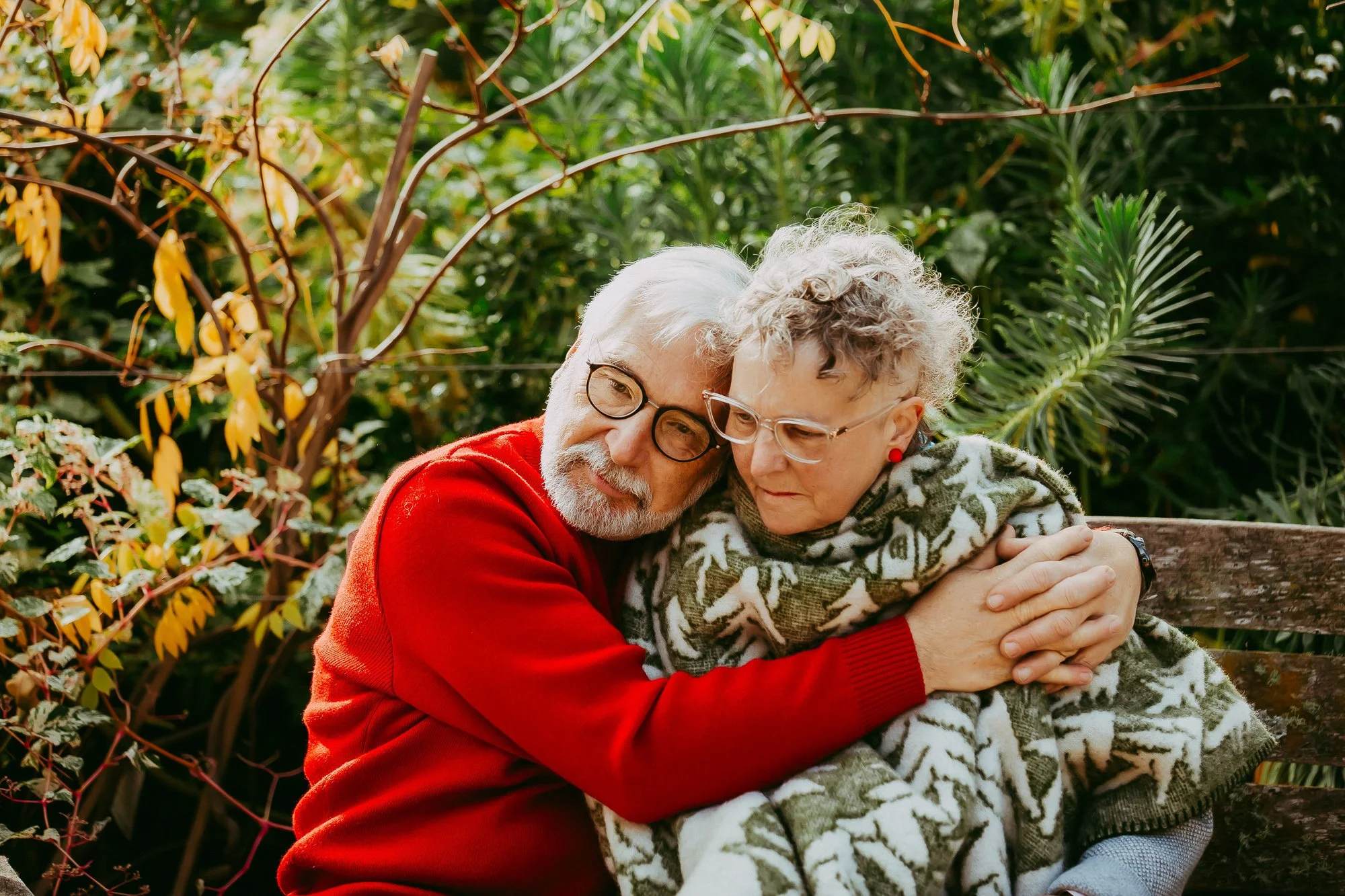 Elderly couple sitting on a wooden bench outdoors, hugging, surrounded by green plants and trees, with the man in a red sweater and the woman in a patterned wool sweater.