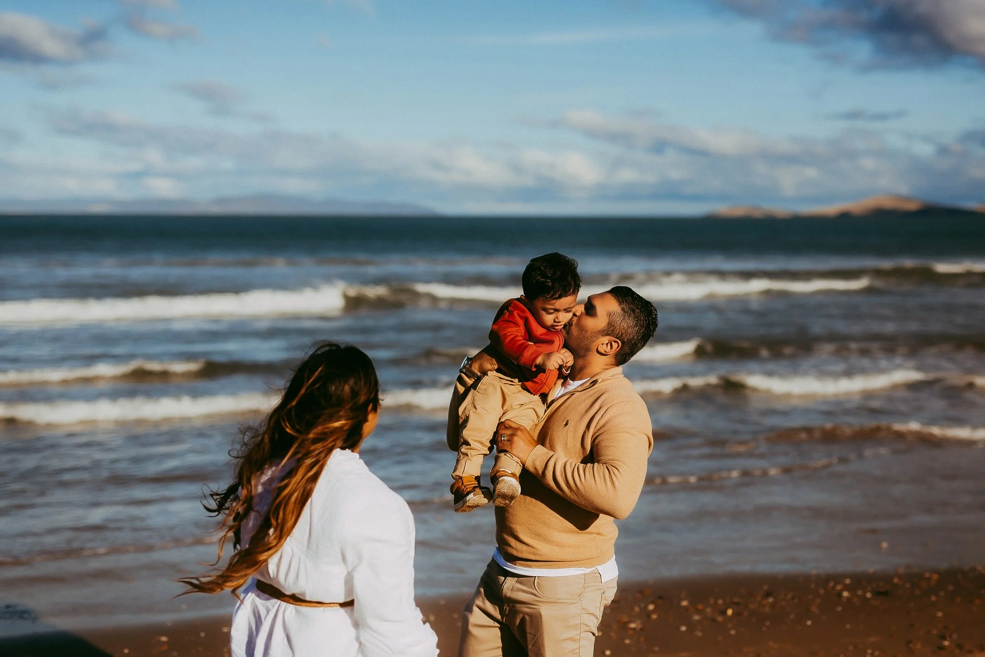 A man holding a young boy in his arms on the beach with an ocean and cloudy sky in the background, a woman with long hair looking at them. Stunning Tasmanian family photo session.