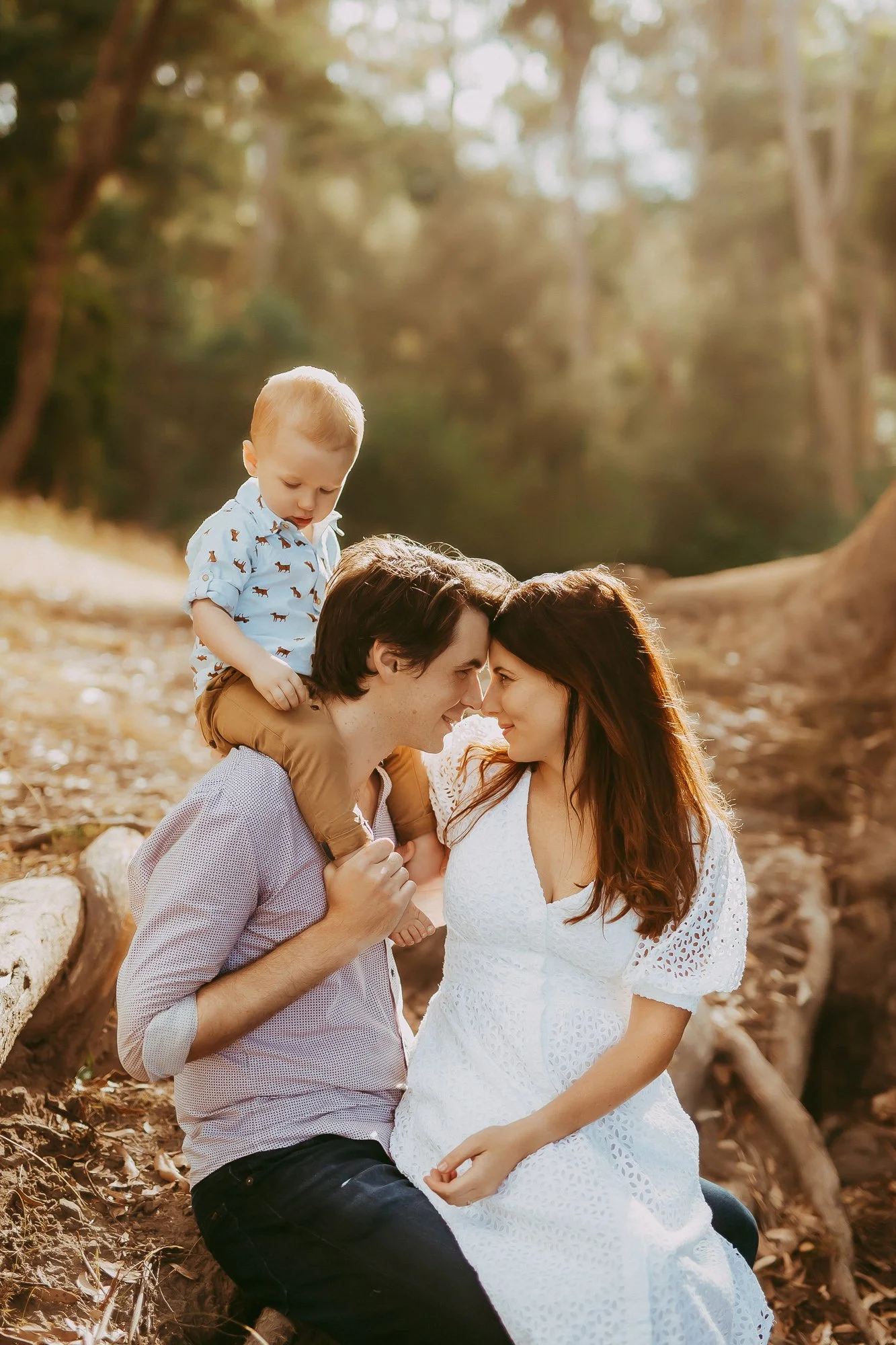 A family of three, with a man, woman, and young boy, sitting on a log outdoors in a wooded area, touching foreheads and smiling at each other, during golden hour.