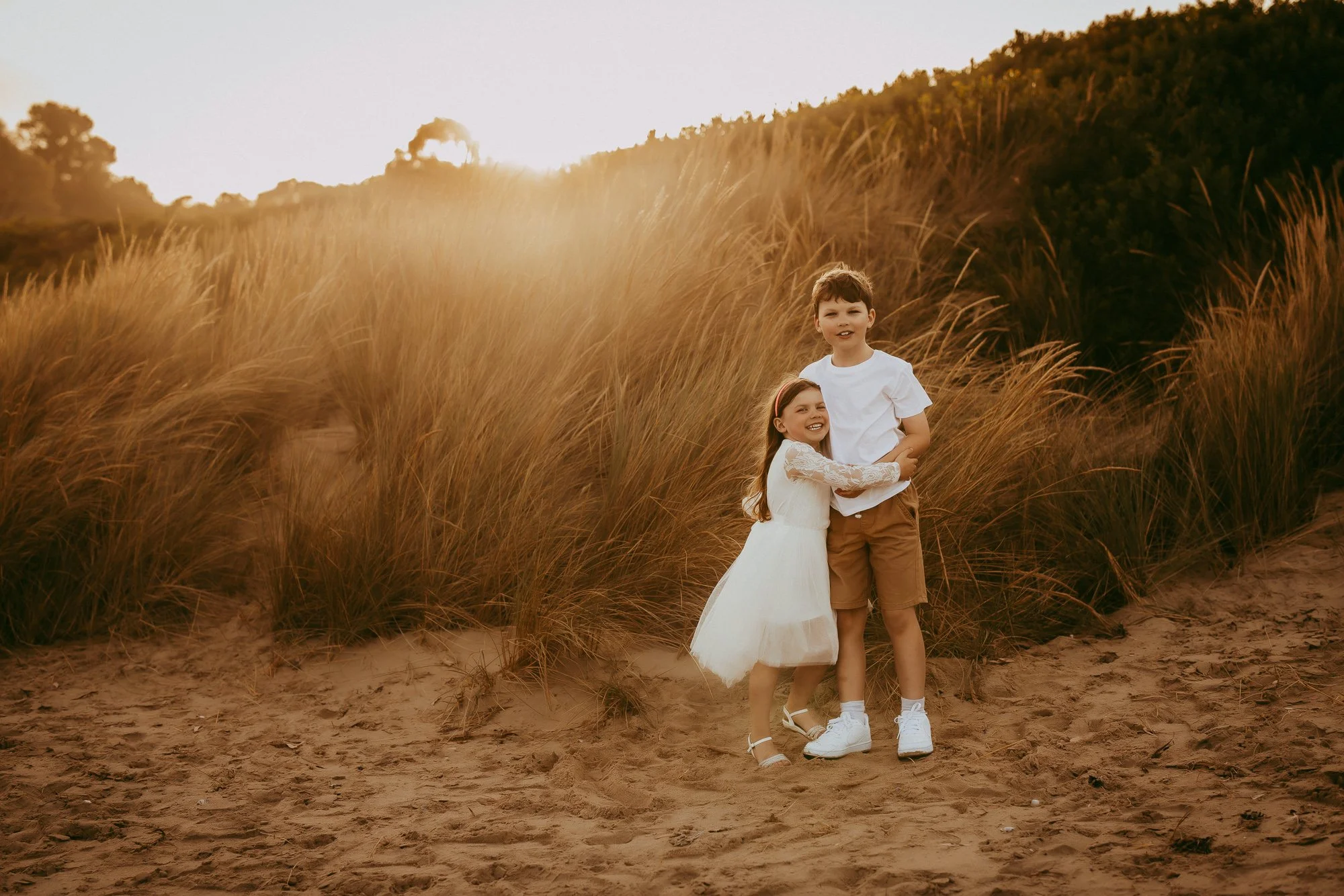 A young boy and girl standing on sandy ground in front of tall grass and bushes at sunset, smiling and hugging each other.