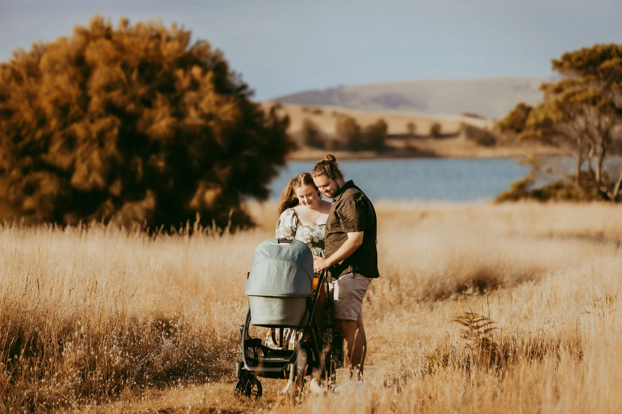 A young couple standing in a golden field with a stroller, looking at it happily, near a lake with trees and hills in the background during a sunny day. South Arm Tasmania family photo session with relaxed approach.
