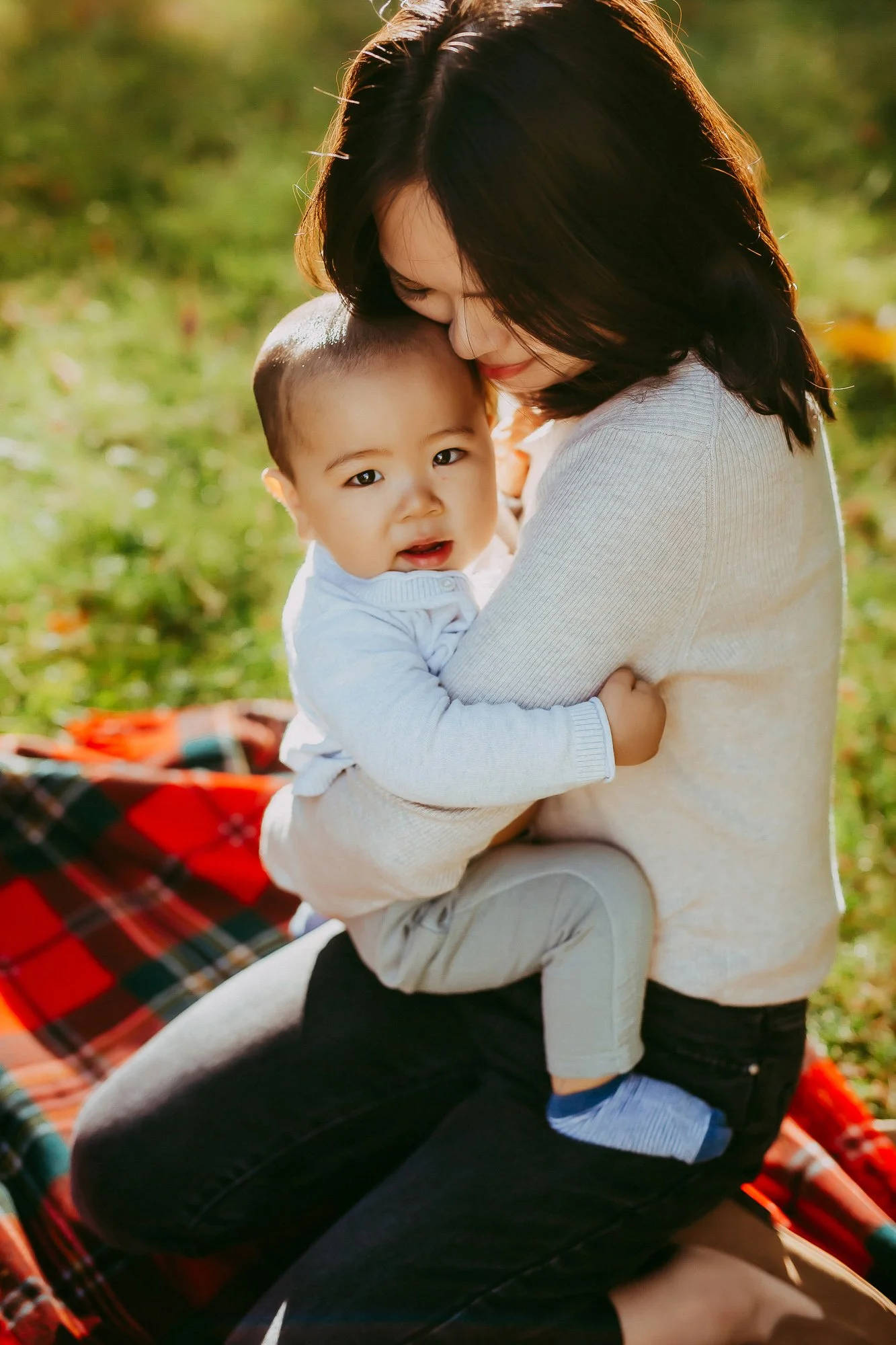 A woman gently holding a young child with short hair, outside on a sunny day, sitting on a red and green checkered blanket in a grassy area. Unposed Hobart family photography by Ulla.