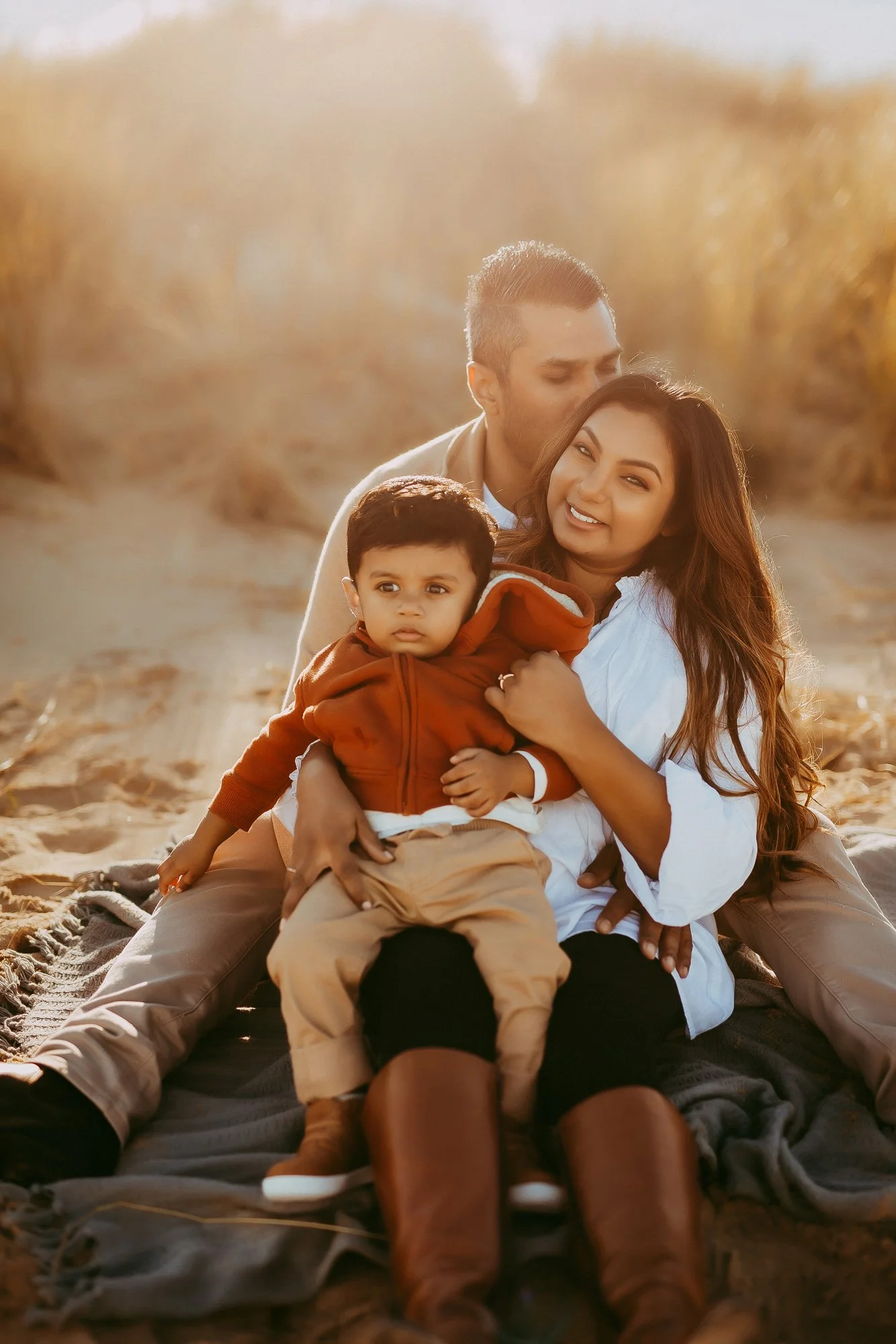 A family of three sitting on a blanket outdoors during sunset, with the father kissing the mother on the temple, the mother smiling at the camera, and a young boy sitting on her lap looking at the camera. Tasmanian Beach family photosession.