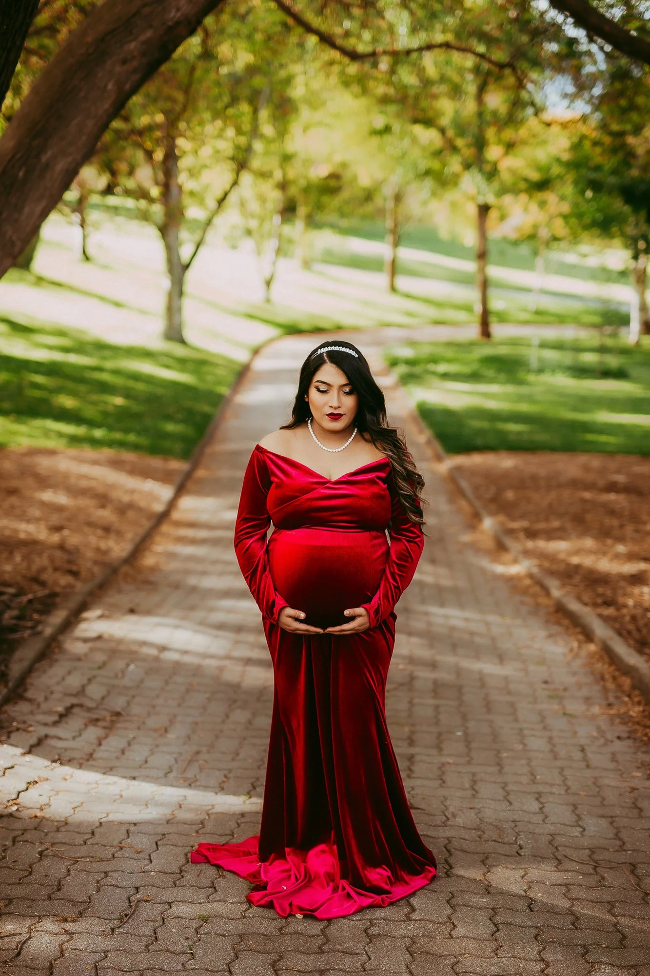 A pregnant woman in a red velvet gown standing on a tree-lined park pathway, gently resting her hands on her belly. She is wearing a pearl necklace and headband, with dark hair styled in loose waves, and is looking down at her belly.