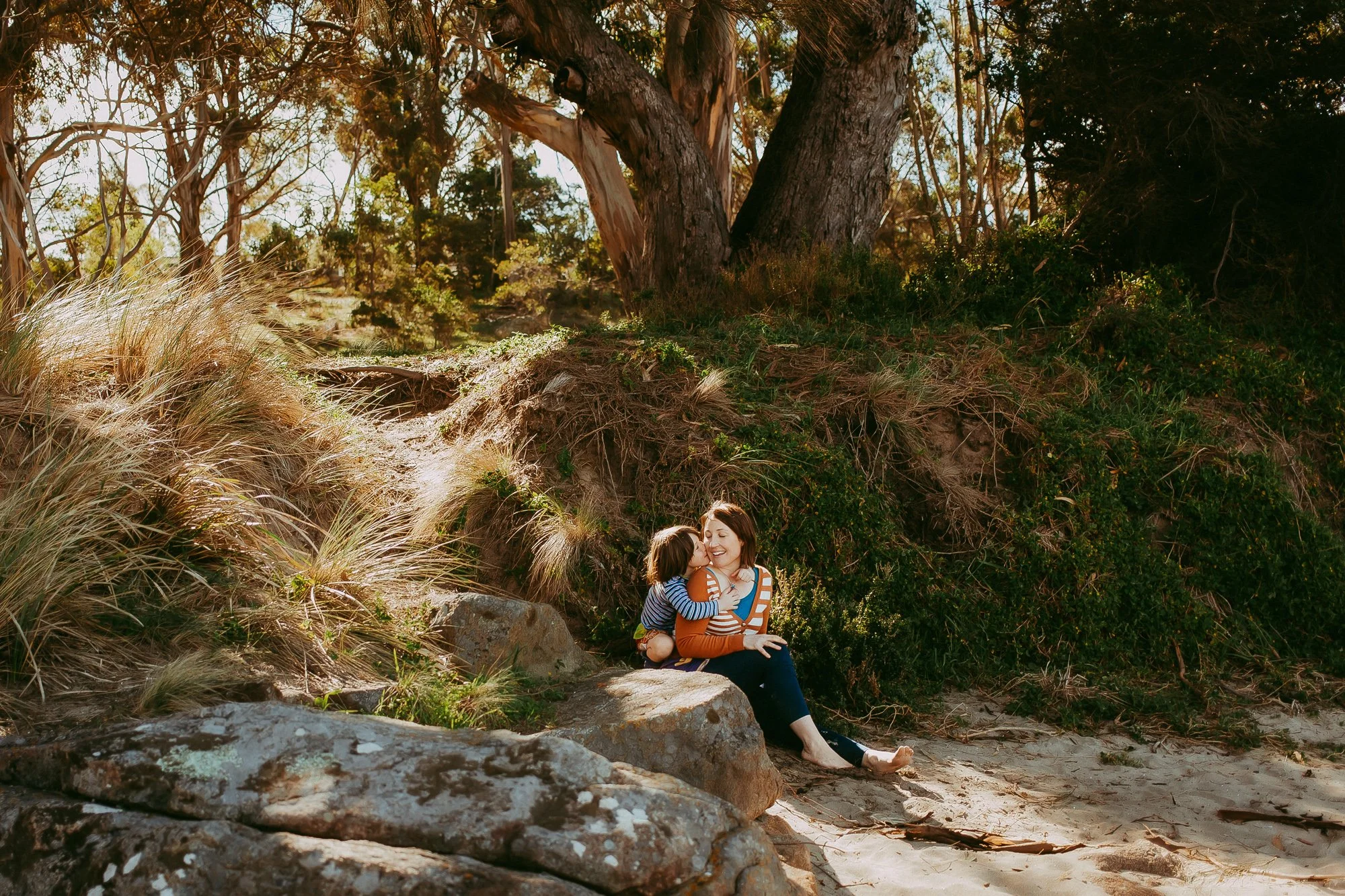Woman and child sitting on rocks at a beach, sharing a kiss with trees and grass in the background. Unposed Hobart family photography.