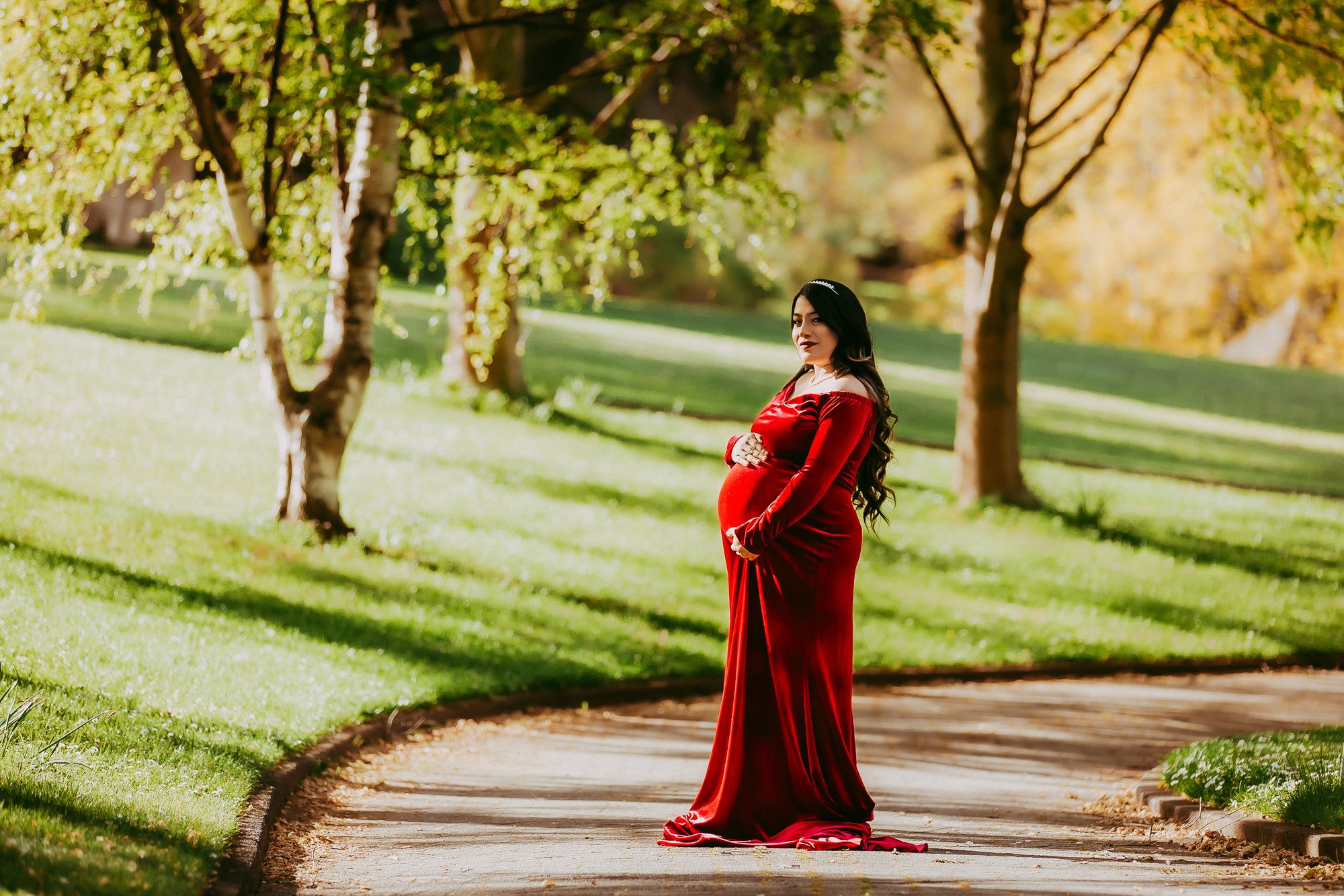 Pregnant woman in a long red dress standing on a park path surrounded by green trees and grass.