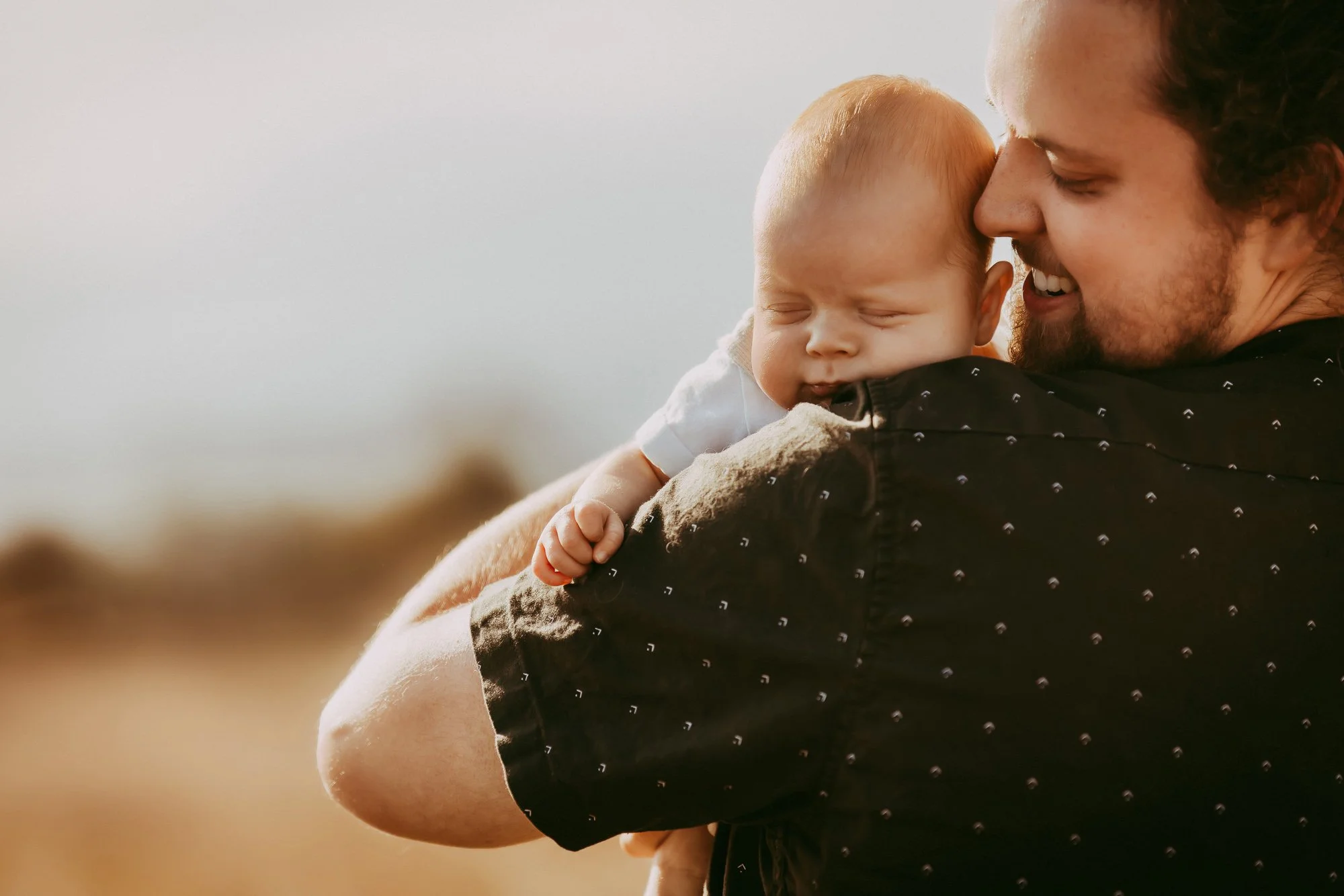 A man holding a sleeping baby outside during sunset, with a blurred natural landscape background. Unposed Hobart family photography with perfect golden hour sunlight.