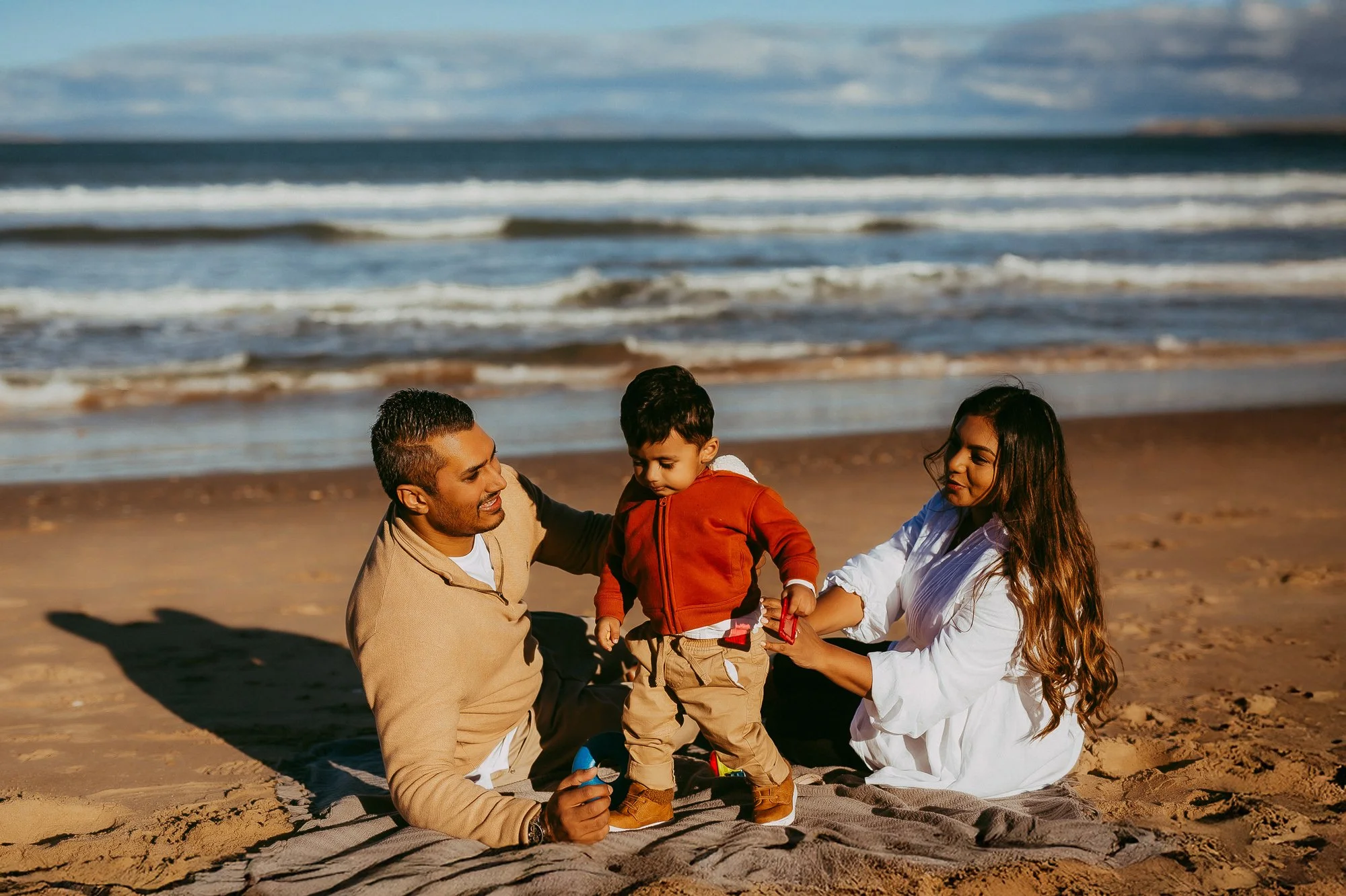 A family of three sits on a blanket on the beach with the ocean and waves in the background, playing with a young boy. Affordable family photographer at Hobart area.
