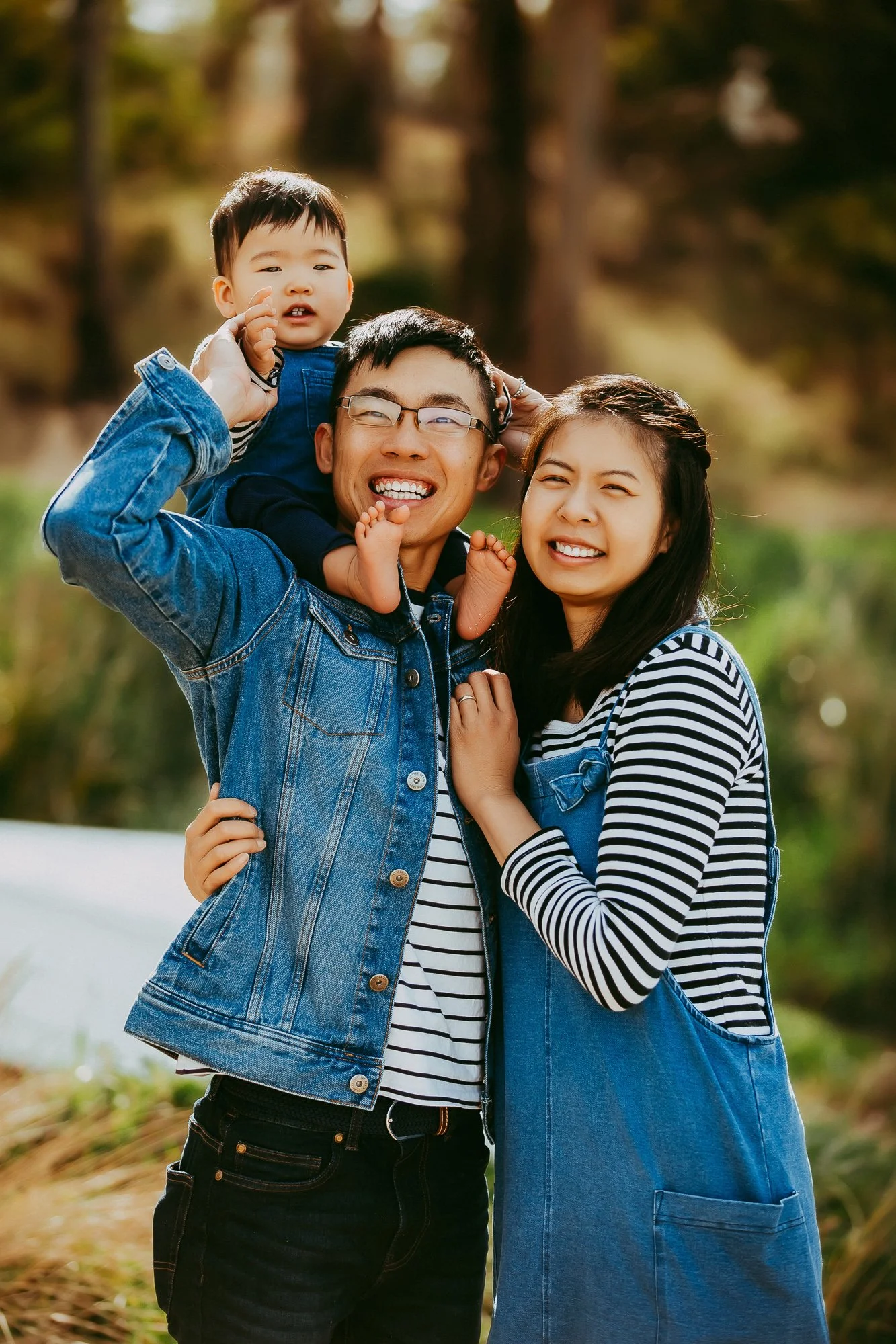 A happy family of three, including a man, woman, and young boy, outdoors in a park with trees in the background, enjoying a playful moment.