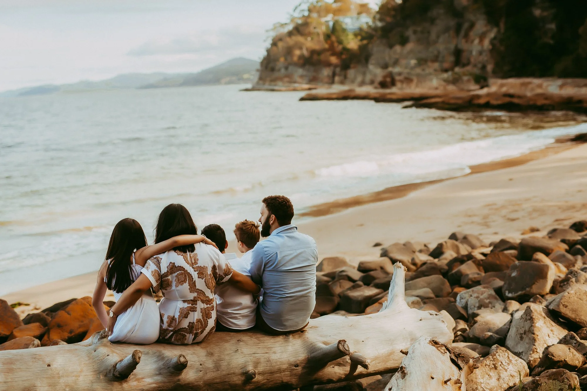 A family of five sitting on a driftwood log on a rocky beach, looking at the ocean and cliffs in the distance.