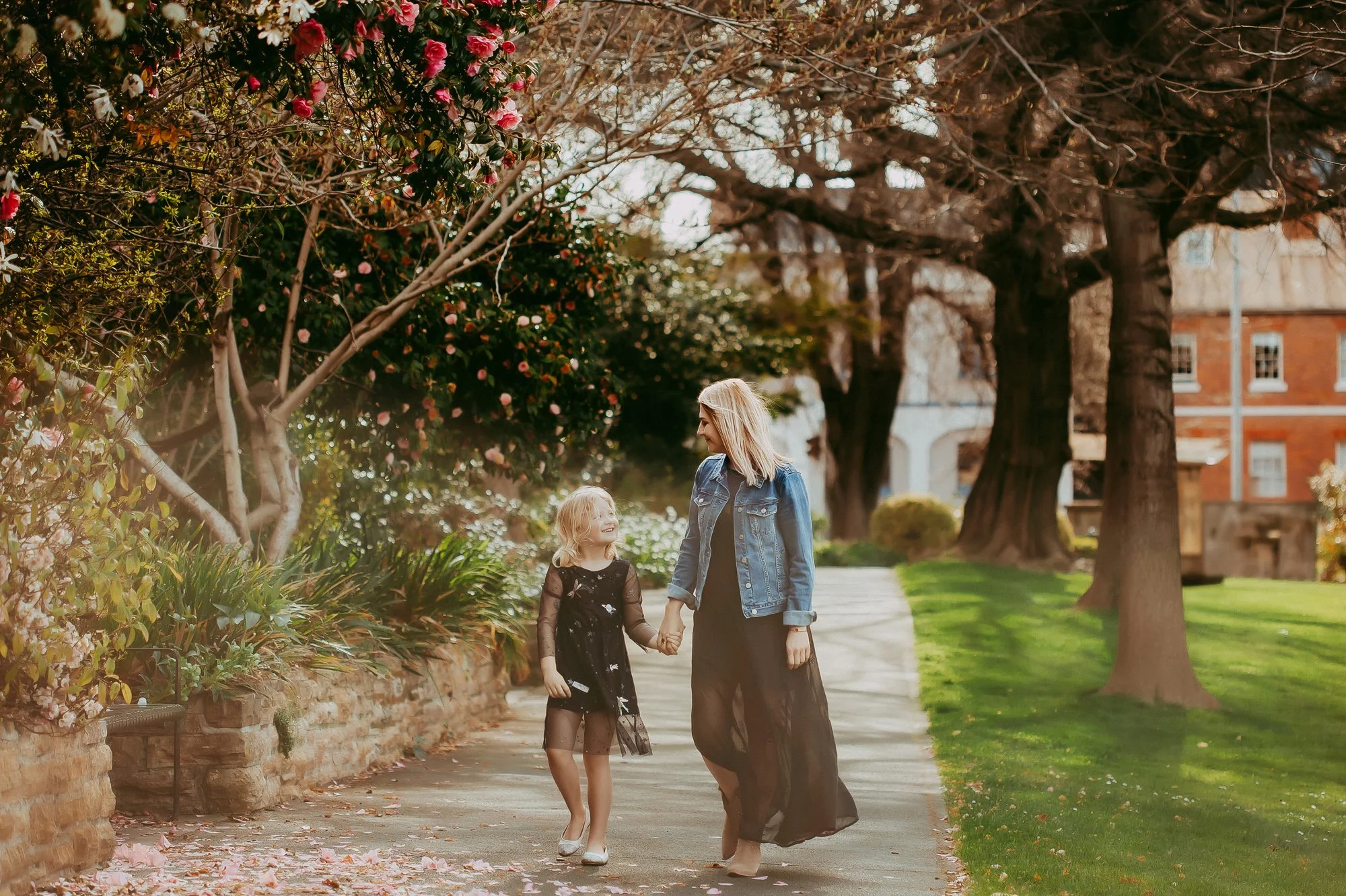A woman and a young girl walking hand in hand on a park sidewalk surrounded by flowering trees and green grass.