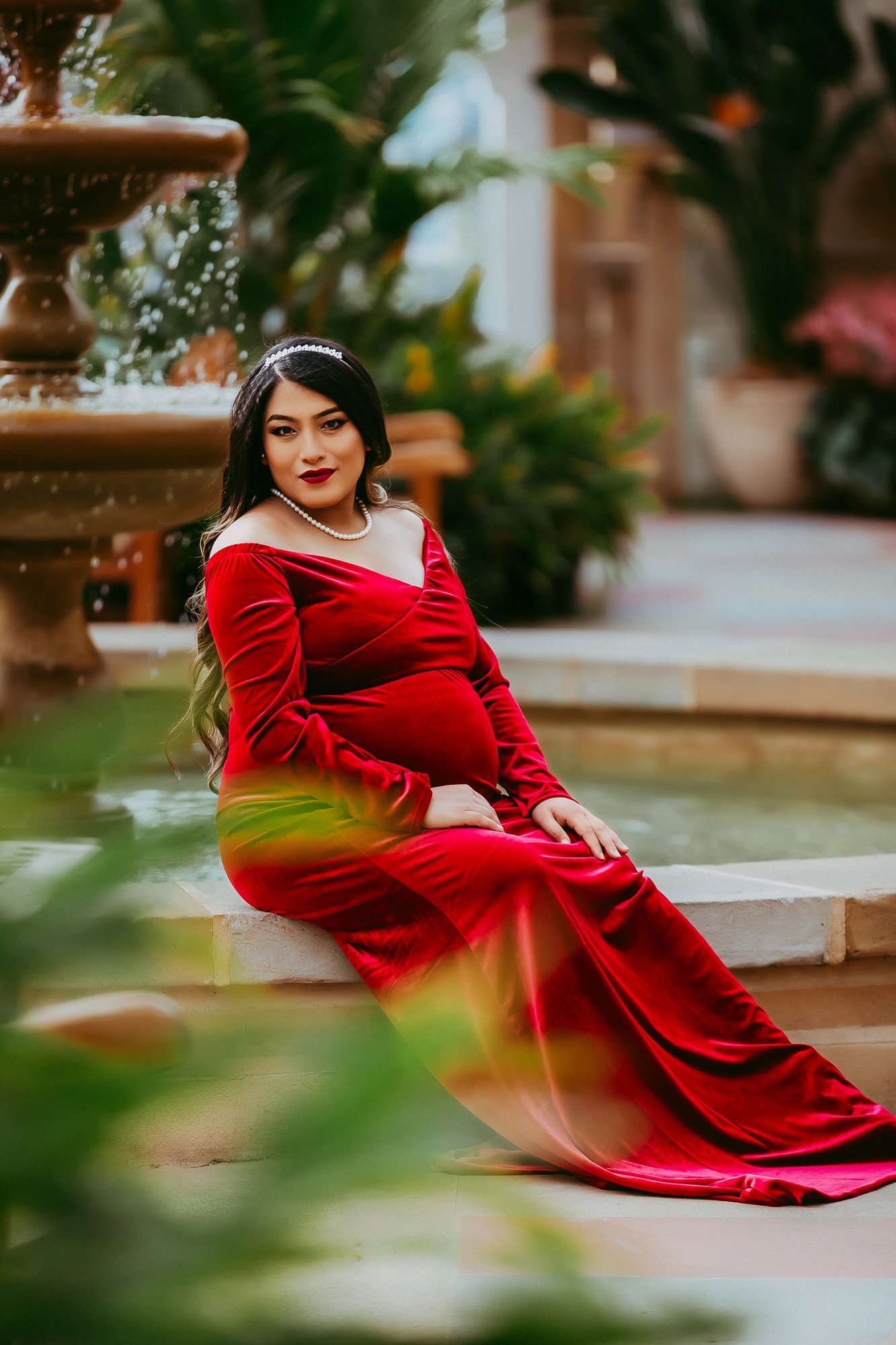 A pregnant woman in a red velvet dress sitting by a fountain outdoors.