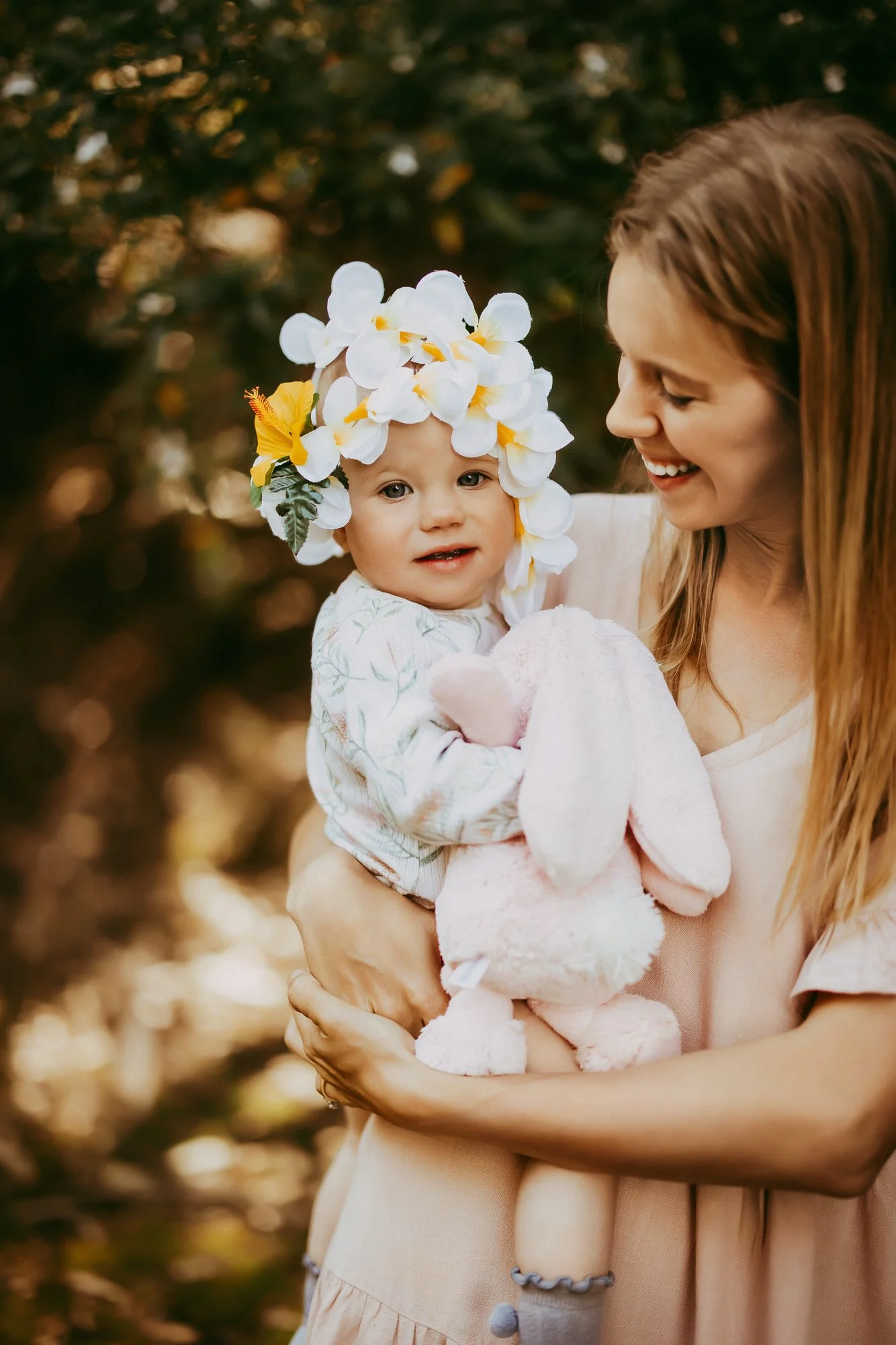 A woman holding a young child outdoors, with the child wearing a flower crown and holding a pink stuffed bunny, smiling at each other.