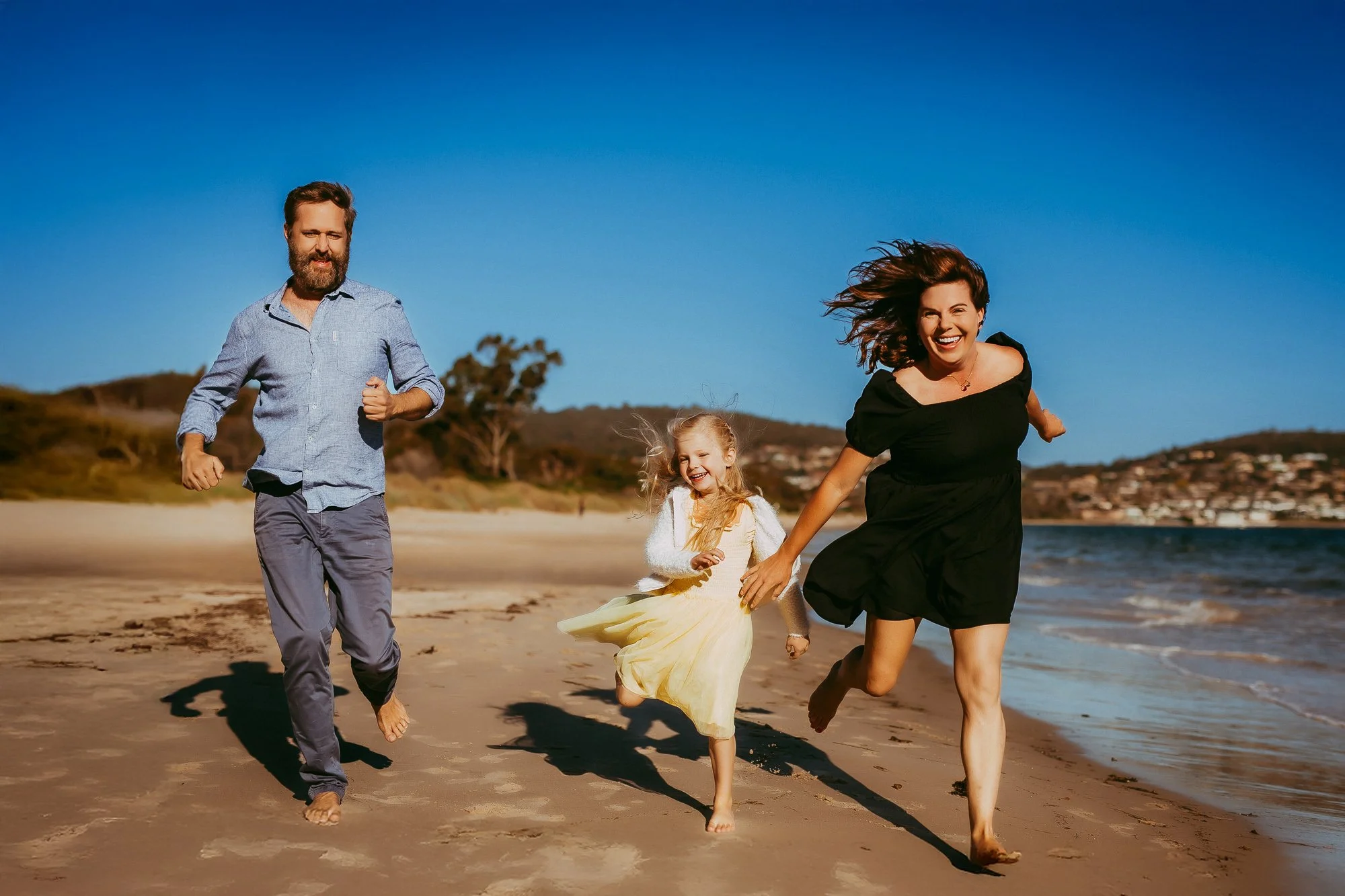 Family of four running barefoot on the beach, smiling and enjoying the sunny day with a clear blue sky and distant houses on a hill in the background. Unposed Hobart family photography by Ulla.