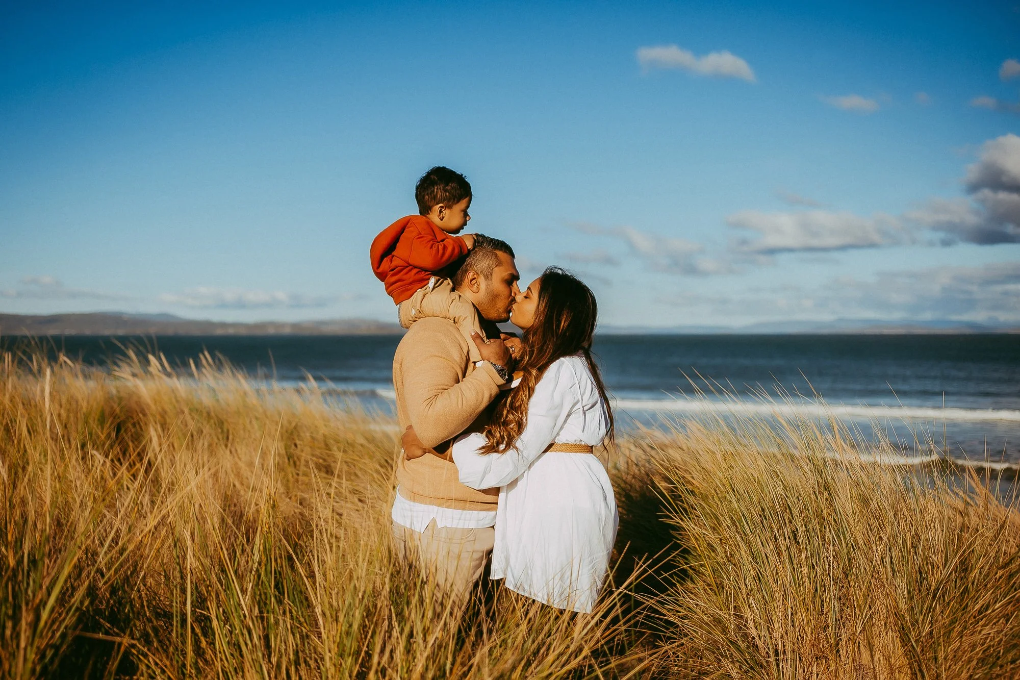A family of three standing in tall grass near the ocean, kissing and embracing, with a blue sky and scattered clouds in the background. Affordable Greater Hobart family photosession.