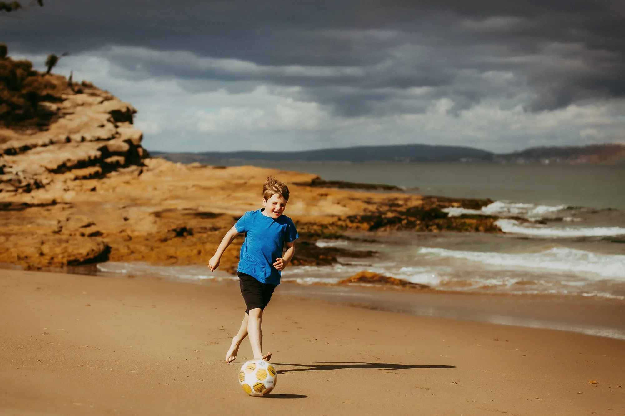 A boy wearing a blue t-shirt and black shorts playing soccer barefoot on a sandy beach with rocky cliffs and an ocean under a cloudy sky.