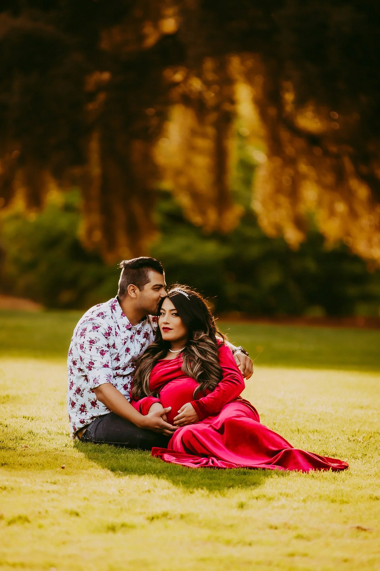 A pregnant woman in a red dress and a man sitting on grass in a park during sunset, with the man kissing the woman's forehead.