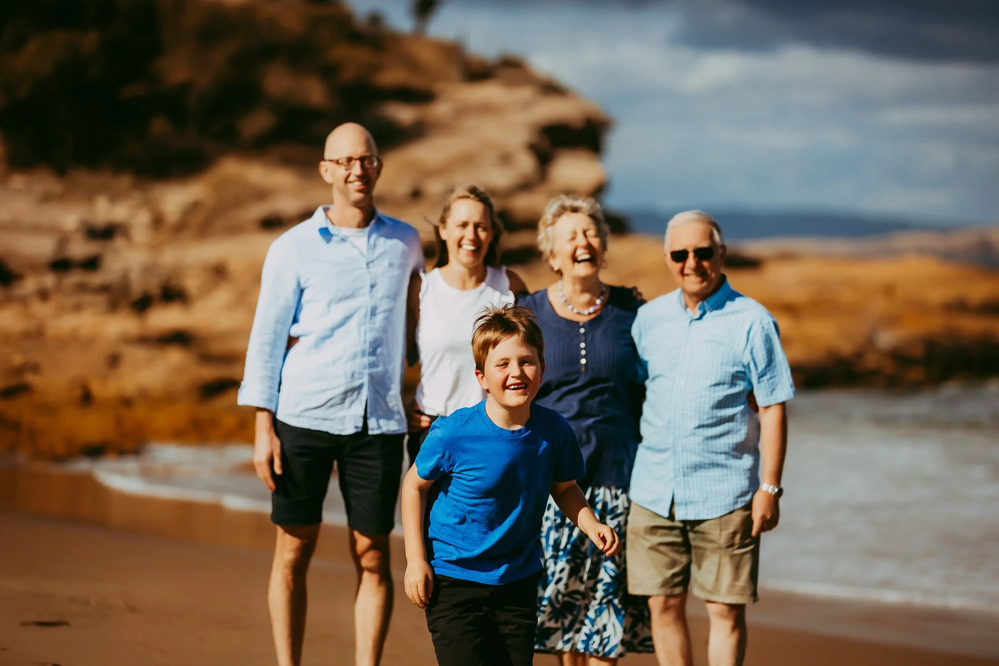 A happy multigenerational family walking along a scenic beach with rocky cliffs in the background, enjoying a sunny day.