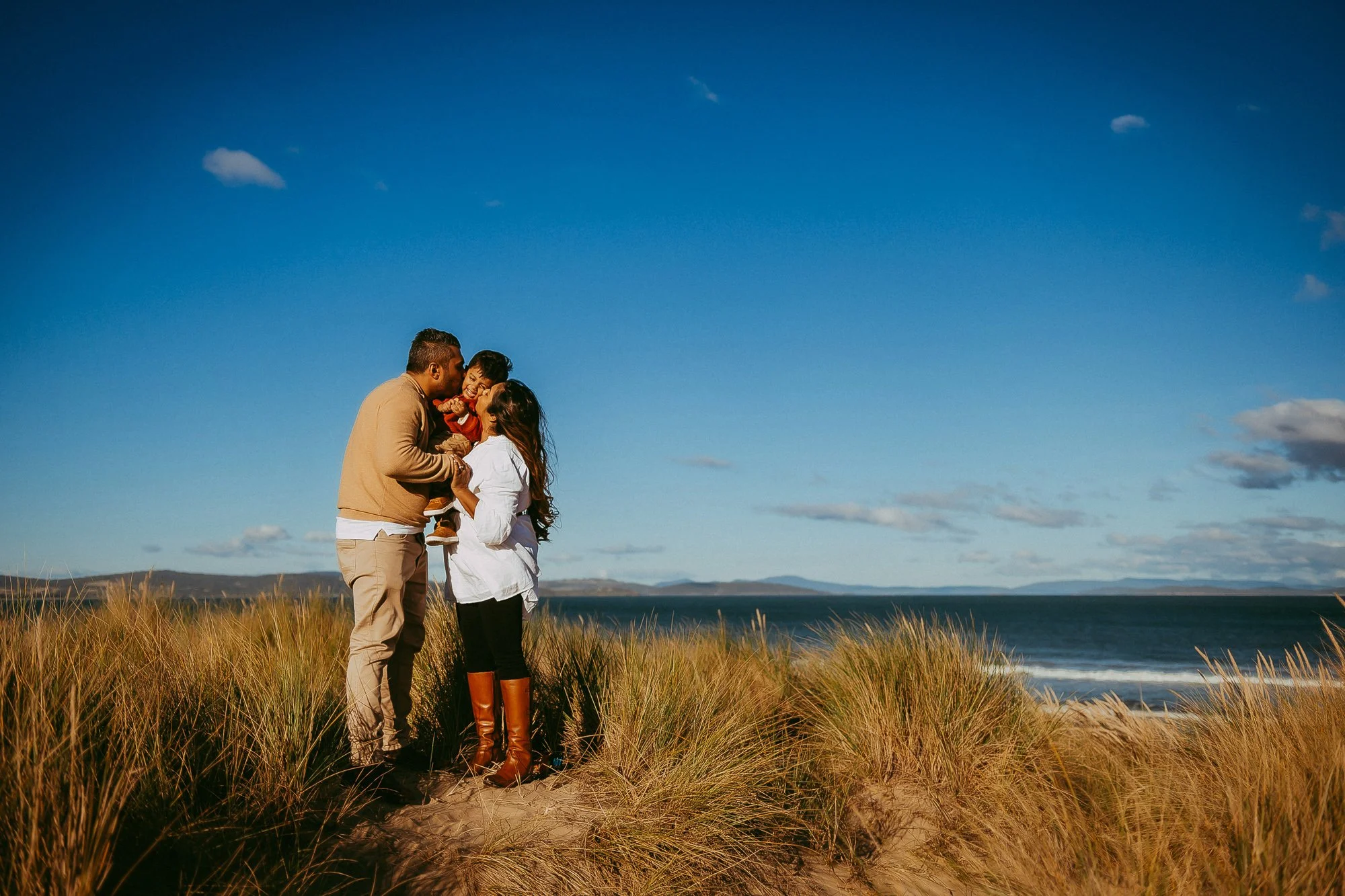 Family of four on a beach with tall grass, a blue sky with scattered clouds, and distant mountains in the background, sharing a joyful moment. Hobart area golden hour family photoshoot.