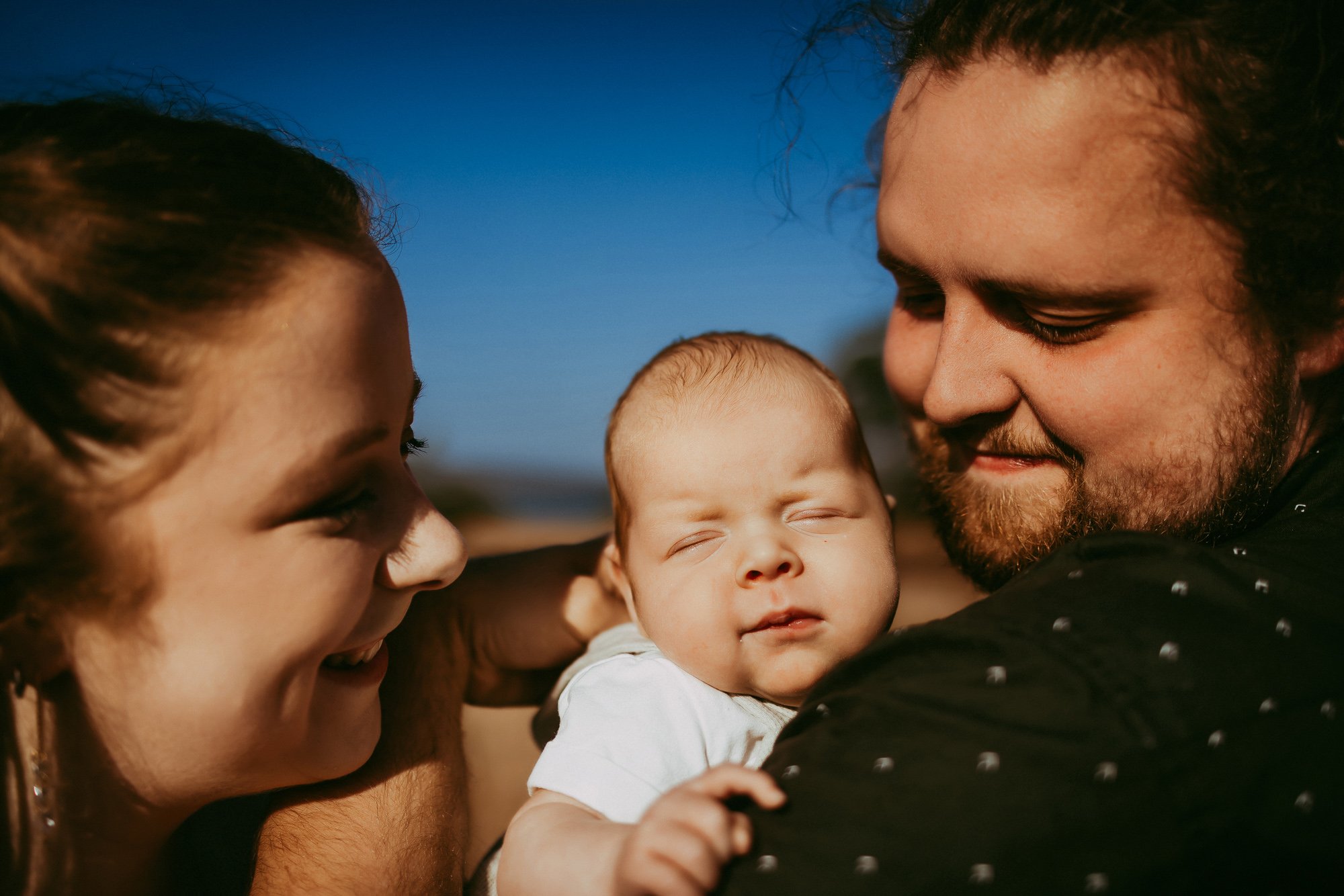 A smiling woman and a man gently hold a sleeping baby outdoors on a sunny day. Seven Mile Beach Tasmania photosession by Ulla, affordable family photographer in Greater Hobart area.