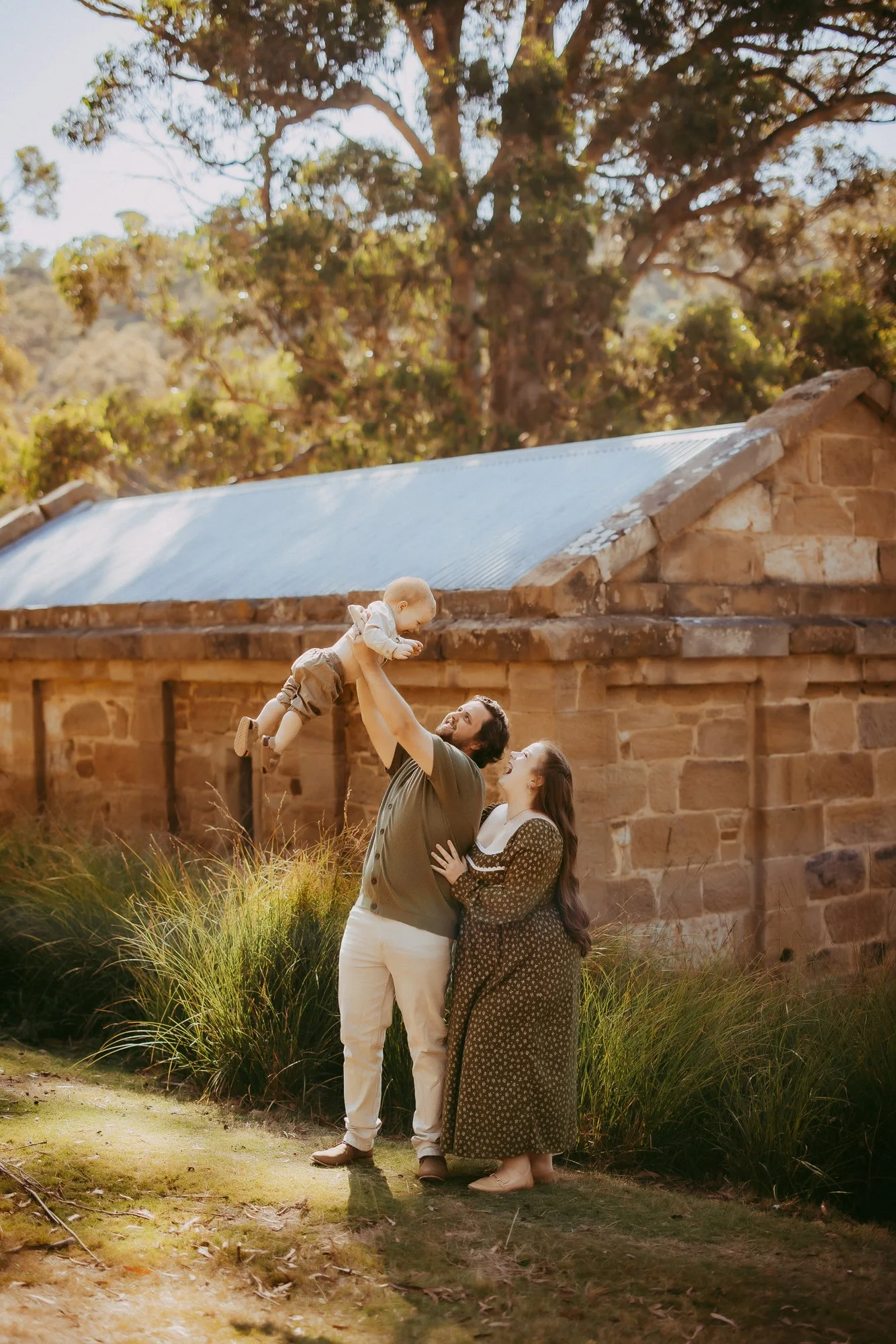 A happy family celebrating outdoors: a man lifts a baby, and a woman stands close, smiling at him, in front of an old brick building on a sunny day. Greater Hobart family photography session.