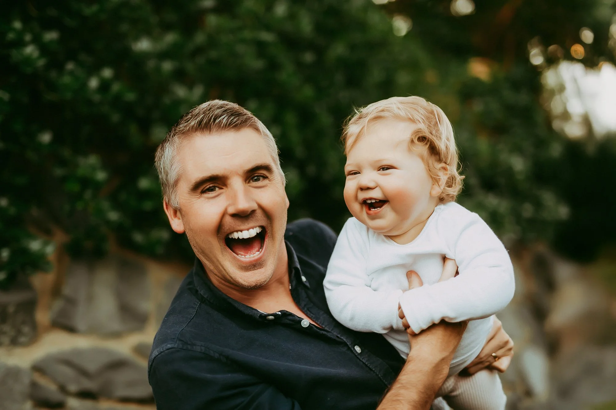 A man holding a smiling young child outdoors in front of a blurred green background. Stunning Tasmanian family photo session.