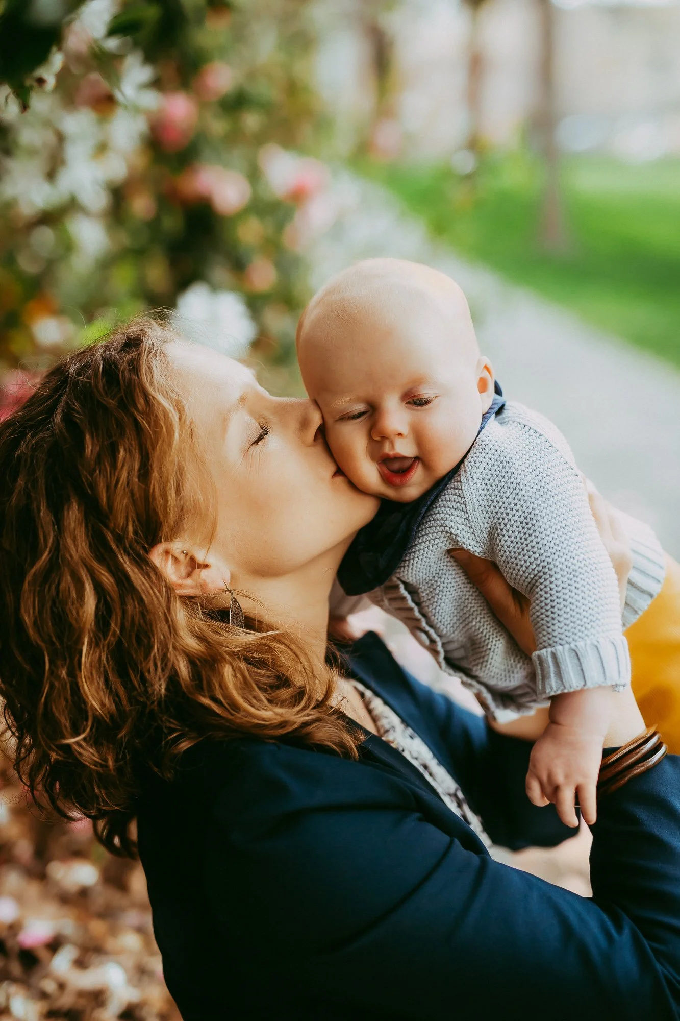 A woman with curly red hair is kissing a baby boy's cheek outdoors, with blurred greenery and flowers in the background.