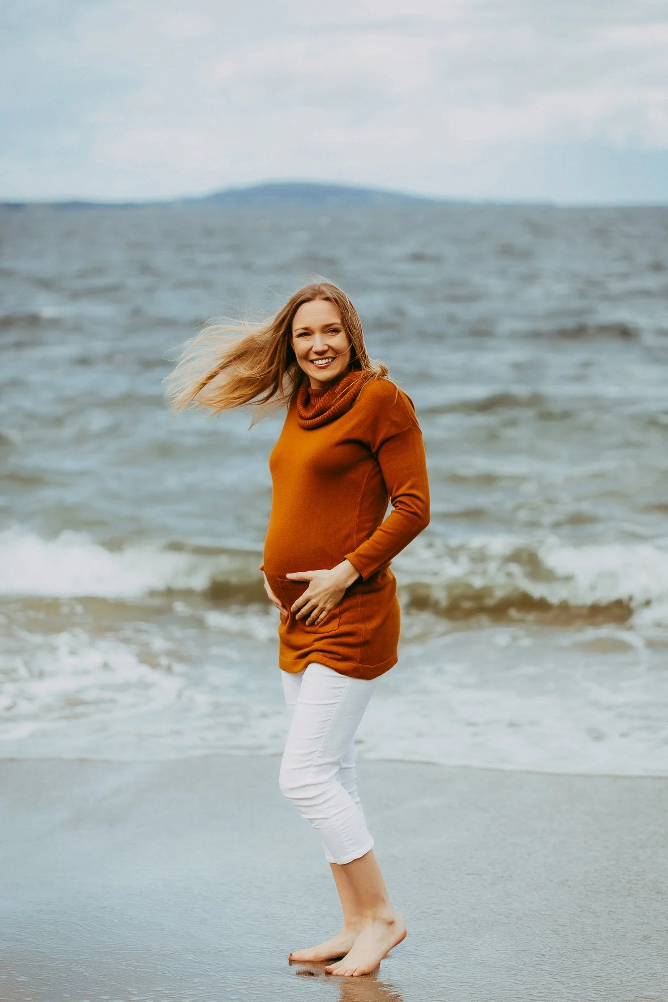 Happy pregnant woman standing barefoot on the beach near the water, smiling at the camera.