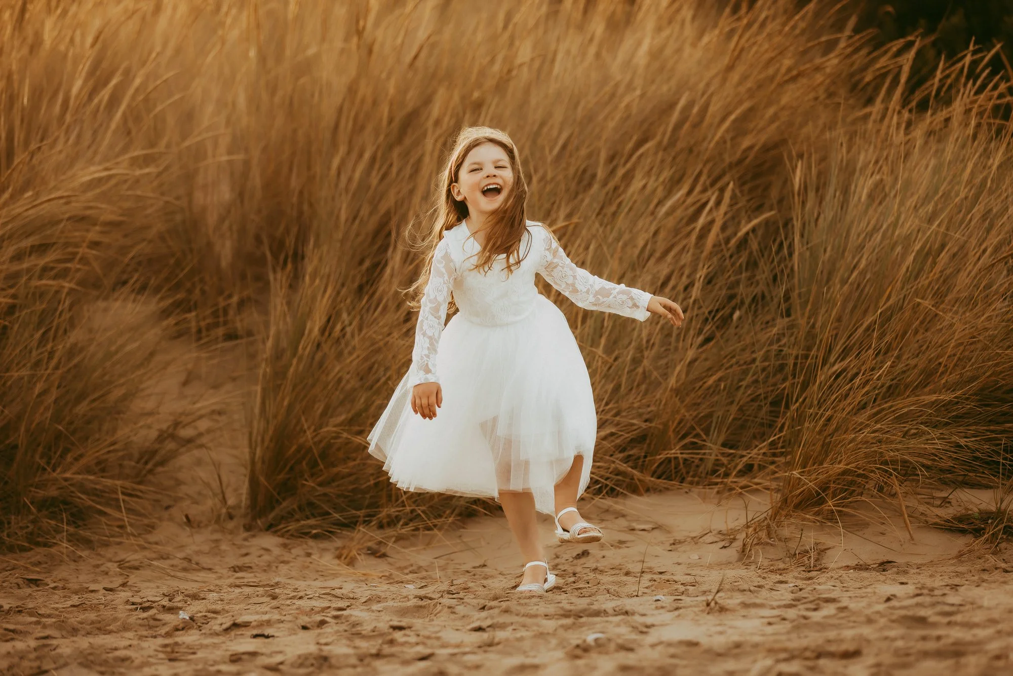 A young girl in a white dress laughing and running through tall, dry grass on a sandy path