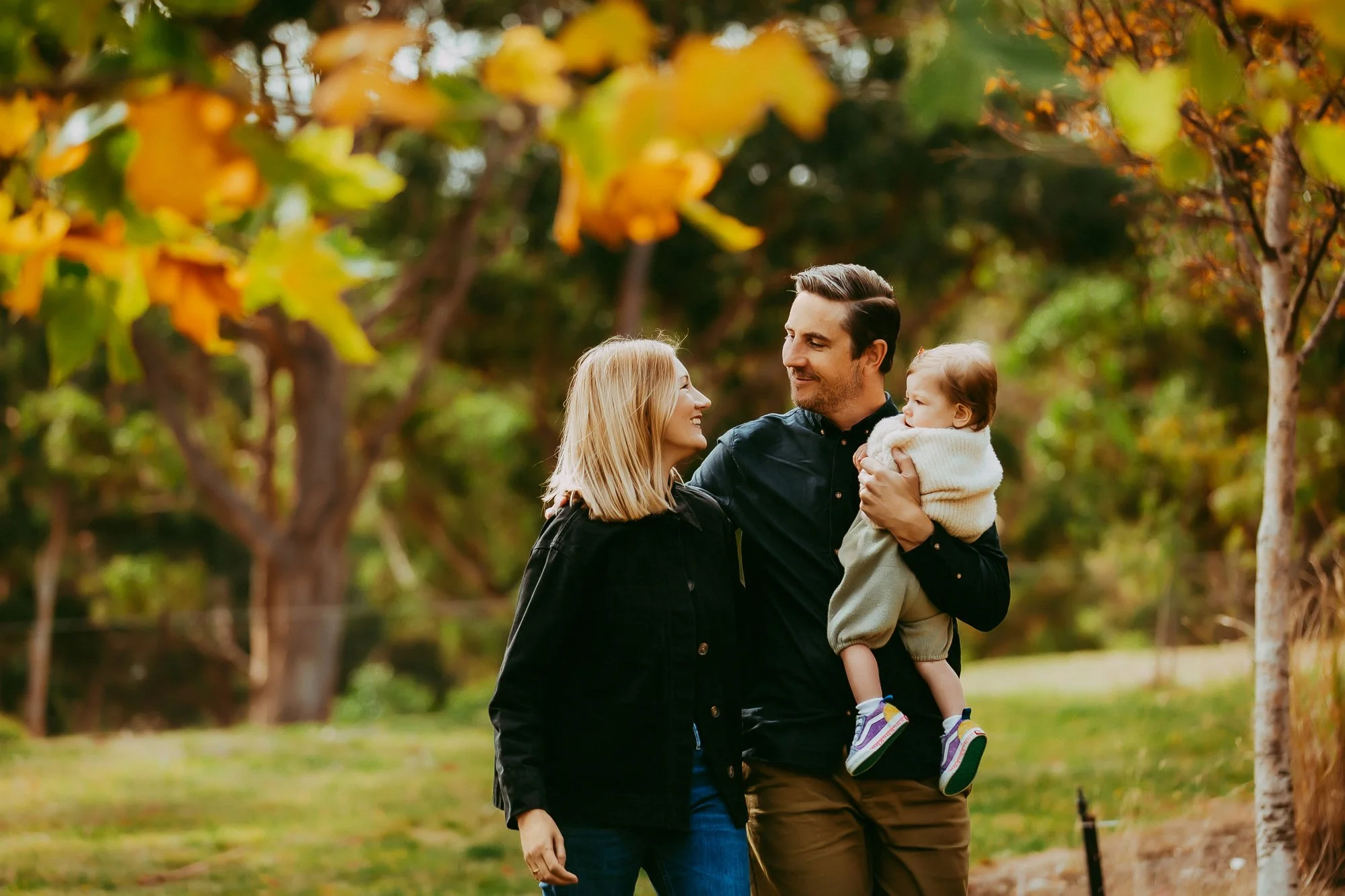 A family of three walking outdoors in an autumn park, smiling at each other. The father is holding a young girl, and the mother walks beside them. Trees with autumn leaves surround them.