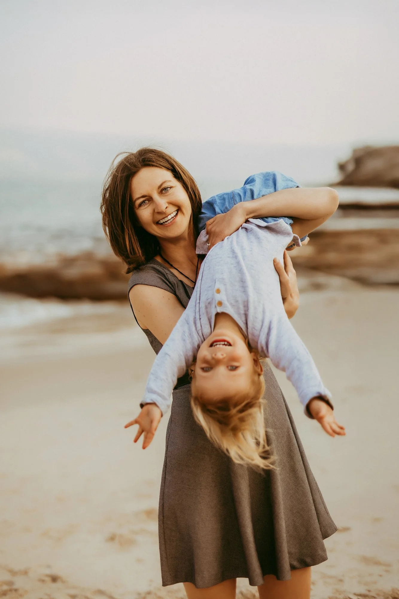 A smiling woman holding a happy blonde girl upside down on a beach, with rocks and the ocean in the background.