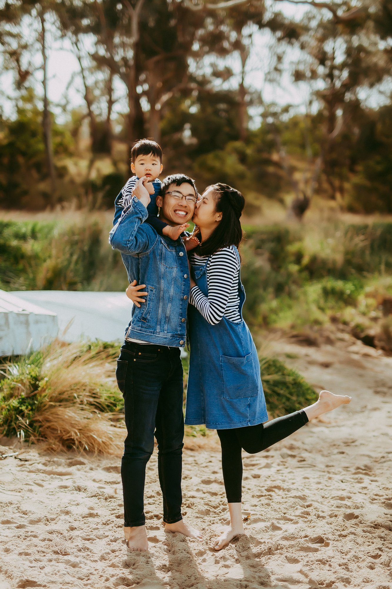 A happy family of three having fun outdoors on a sandy beach. The father is carrying their young son on his shoulder, and the mother is giving the son a kiss. All are smiling and dressed casually, with trees and greenery in the background.