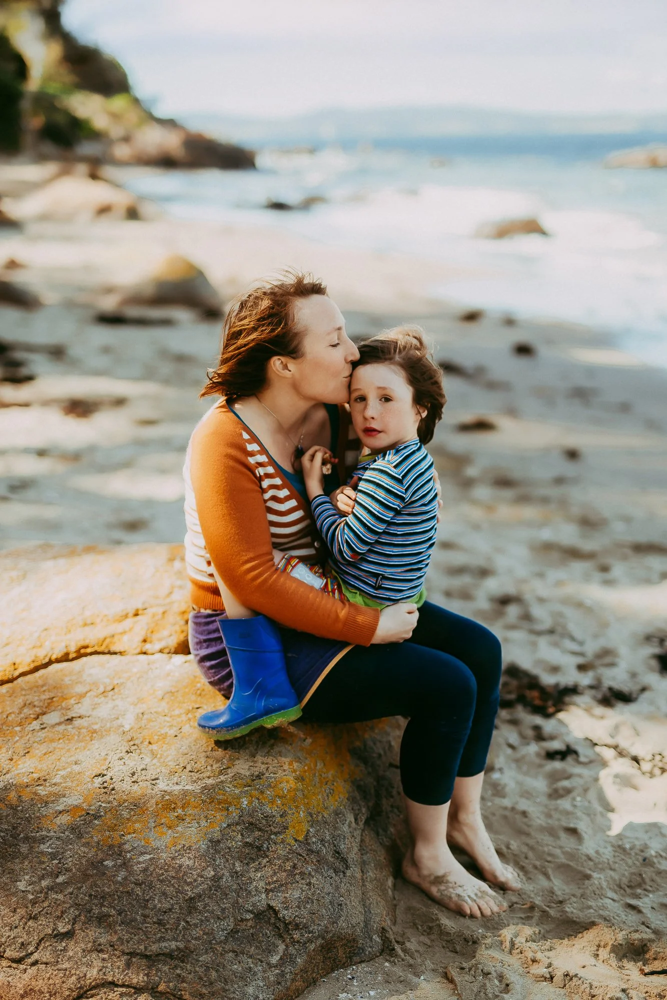 A woman with short brown hair in a striped sweater kisses a young boy with darker hair, wearing a striped shirt, sitting on a large rock at the beach. Tasmanian Beach family photosession.