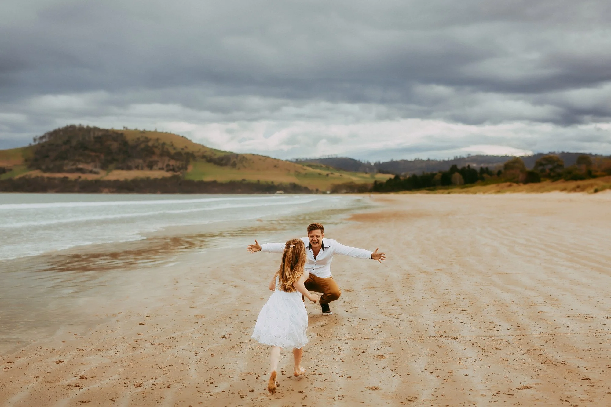 A man and a young girl running and playing on a sandy beach with hills and cloudy sky in the background.