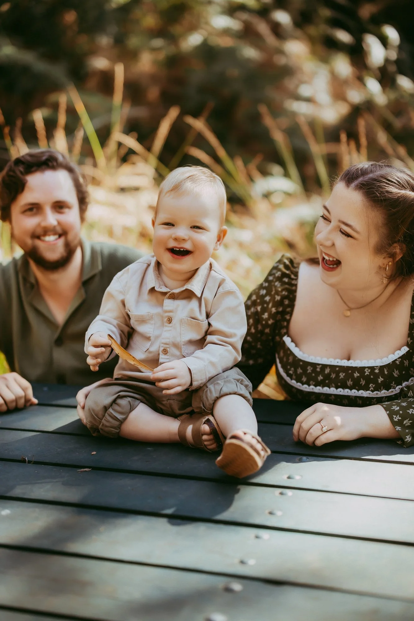 A joyful family with a father, mother, and young child sitting on a wooden picnic table outdoors, smiling and laughing in a natural setting with trees and sunlight in the background. Budget friendly family photosession in Hobart area.