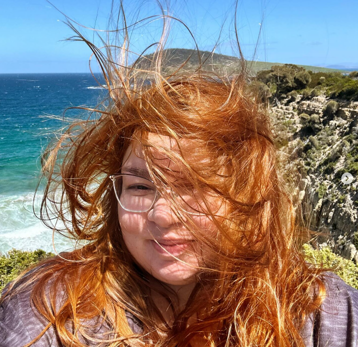 Woman with long red hair, wearing glasses, smiling at the camera on a windy day at the beach with ocean waves and rocky cliffs in the background. Tasmanian beach family photographer.