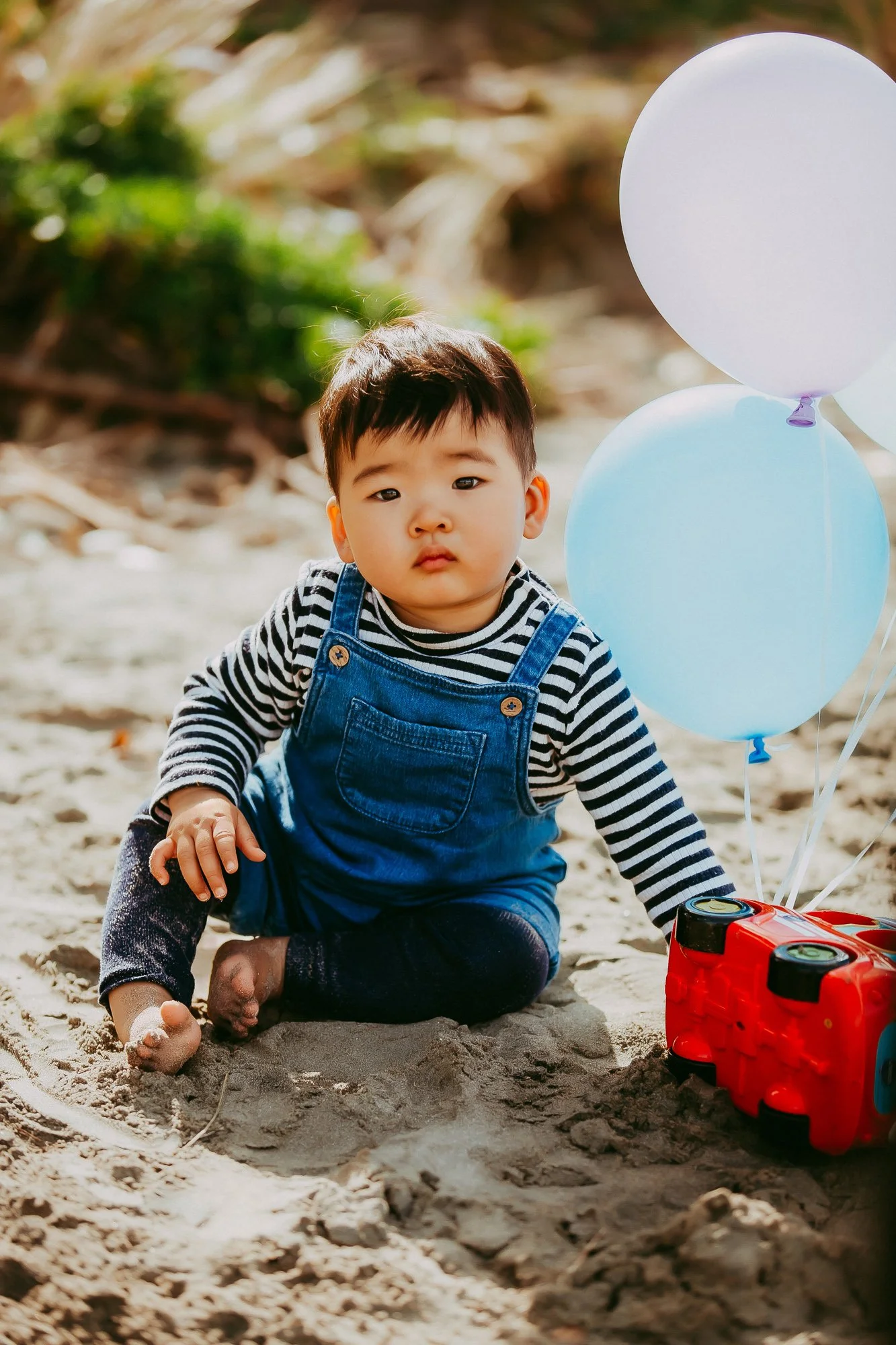 Young Asian boy in striped shirt and denim overalls sitting on the sand at the beach, with balloons and a red toy car beside him.