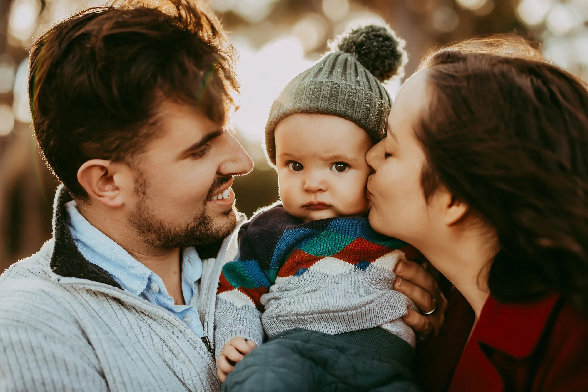 A young family of three outdoors: a man, woman, and baby. The baby wears a knit hat and colorful sweater, the adults smile warmly. Affordable family photoshot.