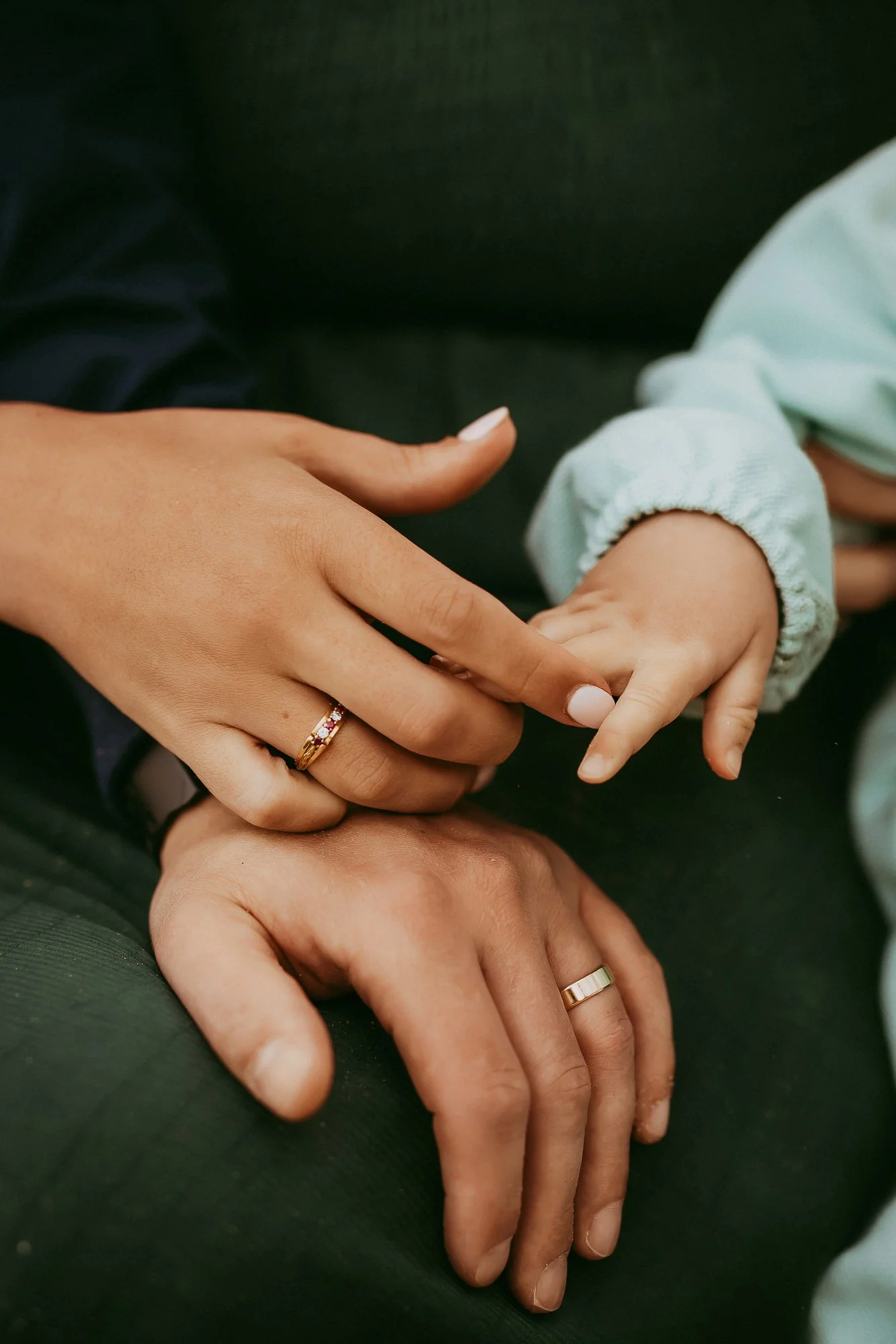 Close-up of a woman and child's hands, both wearing wedding rings. The woman's hand rests on her lap, and she gently holds the child's tiny hand, which is dressed in a light blue sleeve.