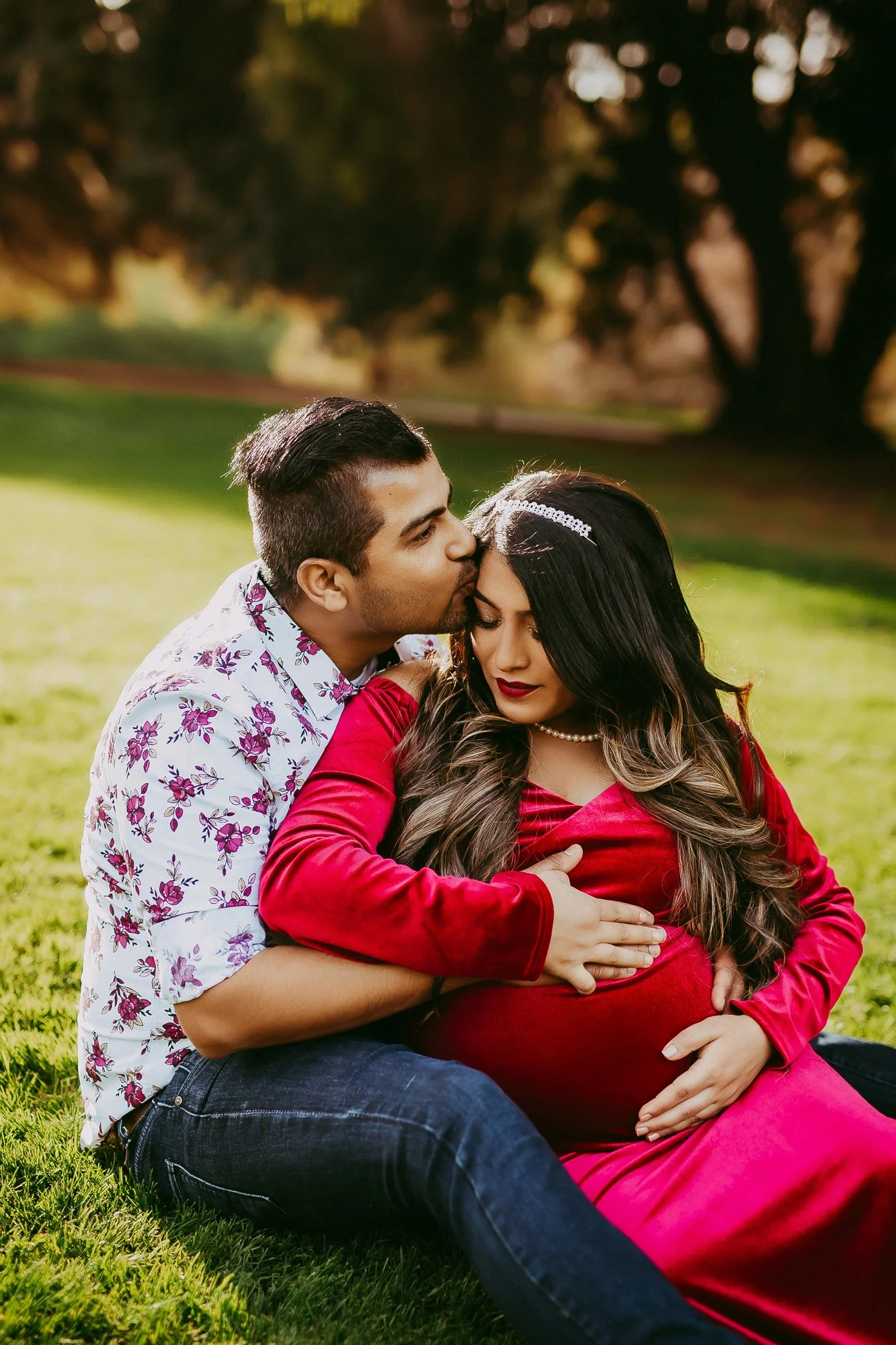 A pregnant woman in a red dress sitting on the grass with a man in a white floral shirt kissing her forehead, outdoors with trees in the background during sunset.
