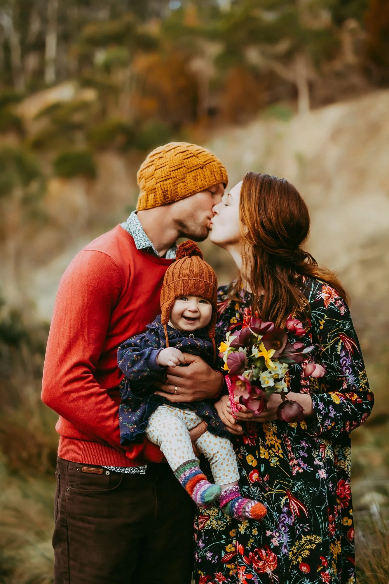 A couple with a young child sharing a kiss outdoors during fall, with the woman holding a bouquet of flowers and the baby smiling, all dressed in warm clothing.