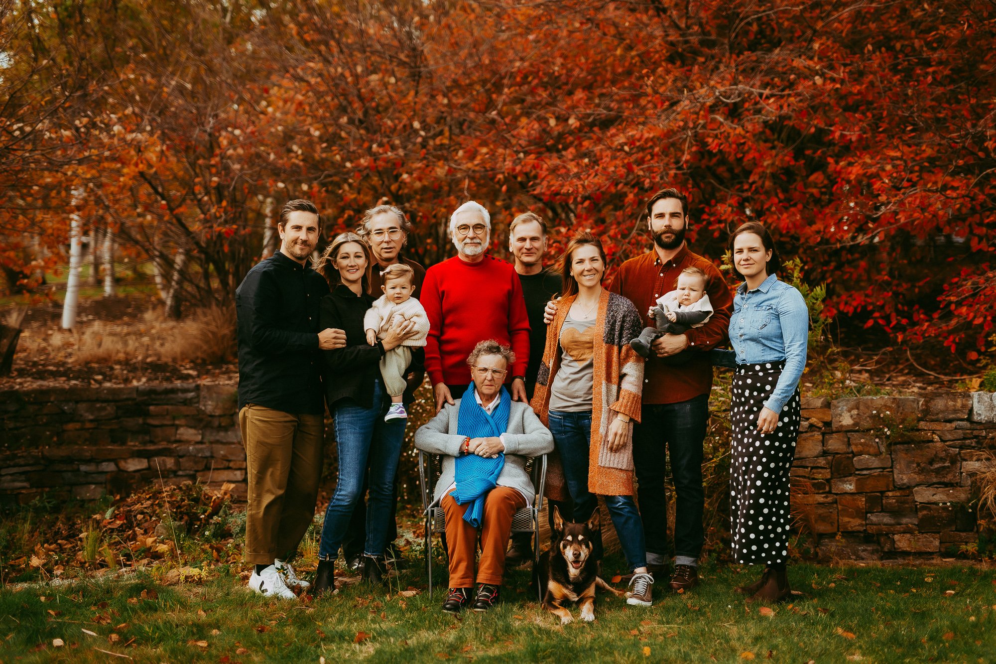 A multigenerational family photo outdoors during autumn, with fall foliage background. Twelve people, including children, adults, and elderly, are standing and sitting, with a dog in front, all smiling.